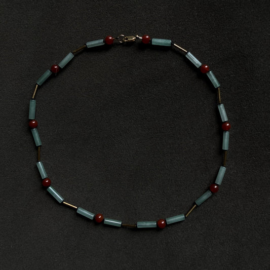 A beaded necklace featuring light blue-green rectangular beads separated by small dark brown cylindrical beads and accented with round dark red beads, displayed on a dark textured background.