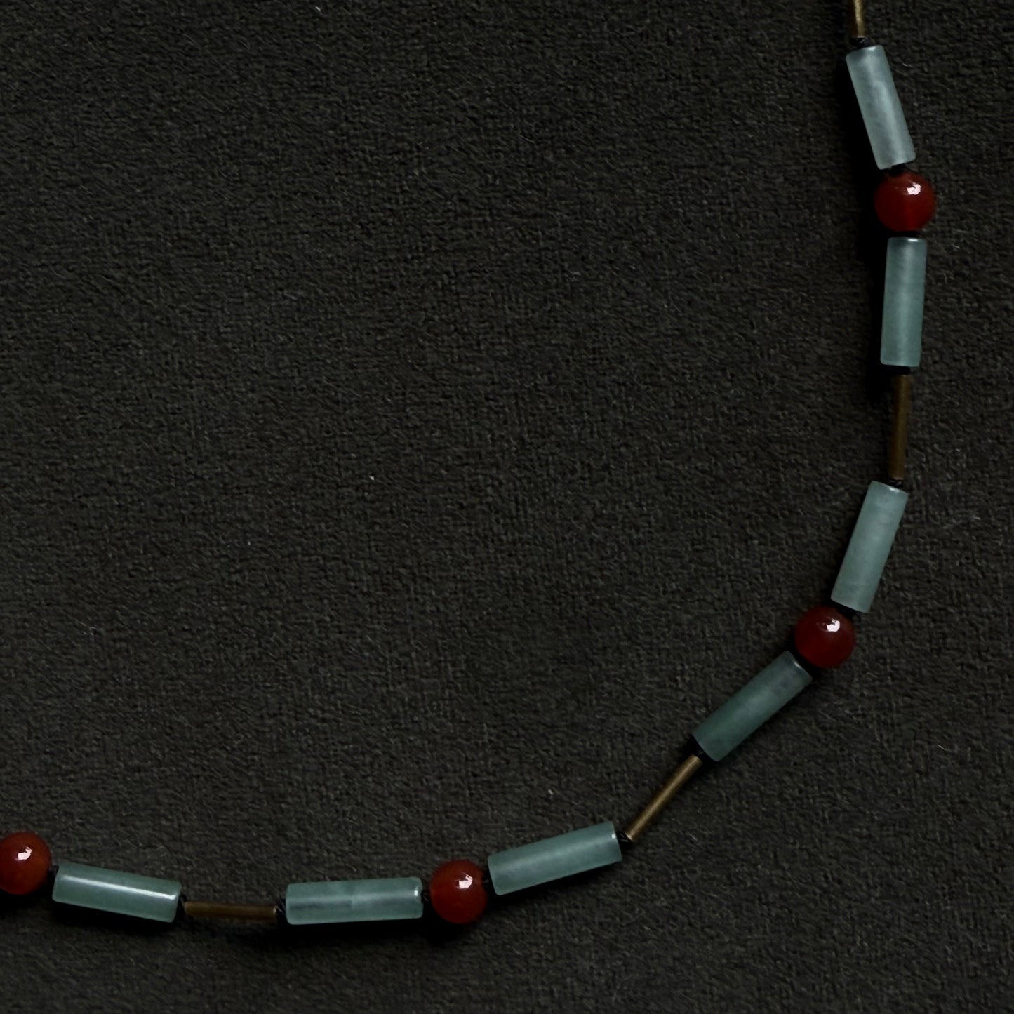 A beaded necklace featuring light blue-gray cylindrical glass beads and small red spherical beads, displayed on a dark textured background.