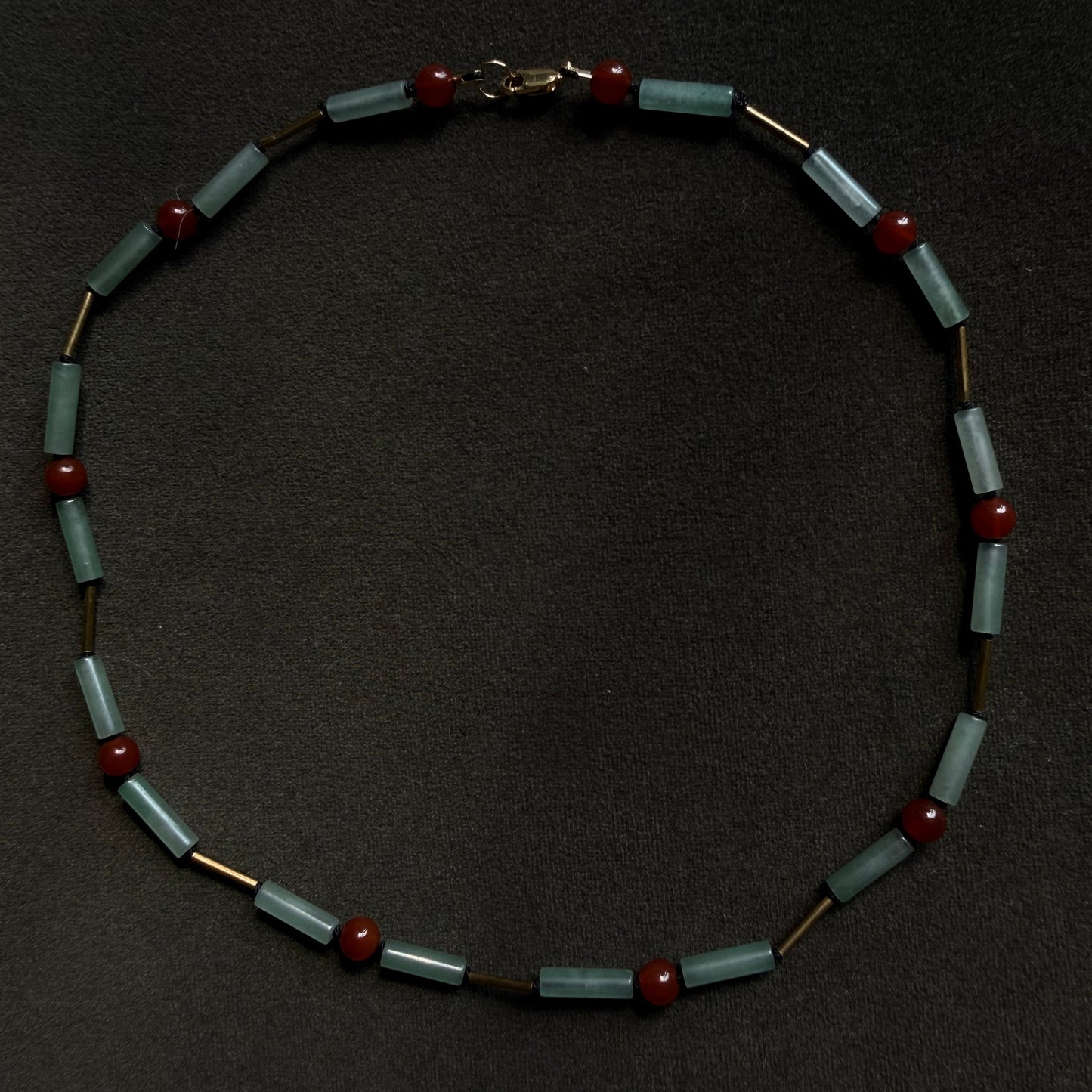A beaded necklace featuring light blue-green rectangular beads, small red round beads, and gold-toned spacer rings, displayed on a dark textured background.
