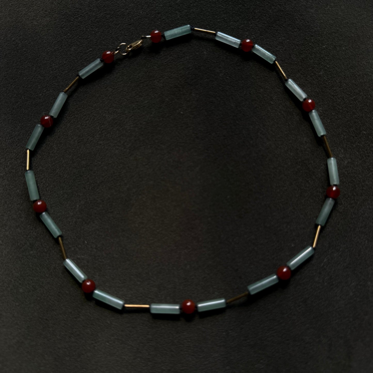 A beaded necklace featuring light blue-gray rectangular beads, small dark red round beads, and gold-toned spacer rings, arranged in a circle on a dark textured background.