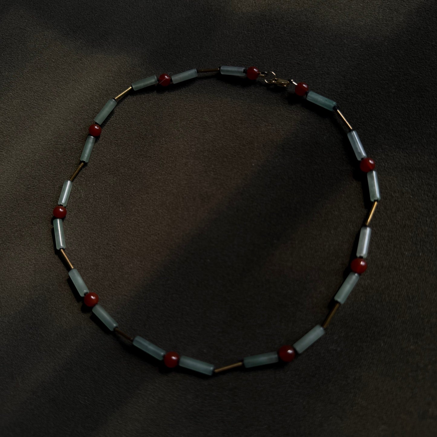 A beaded necklace with alternating light blue-gray rectangular beads and small red round beads, separated by thin gold-toned links, displayed on a dark textured background.