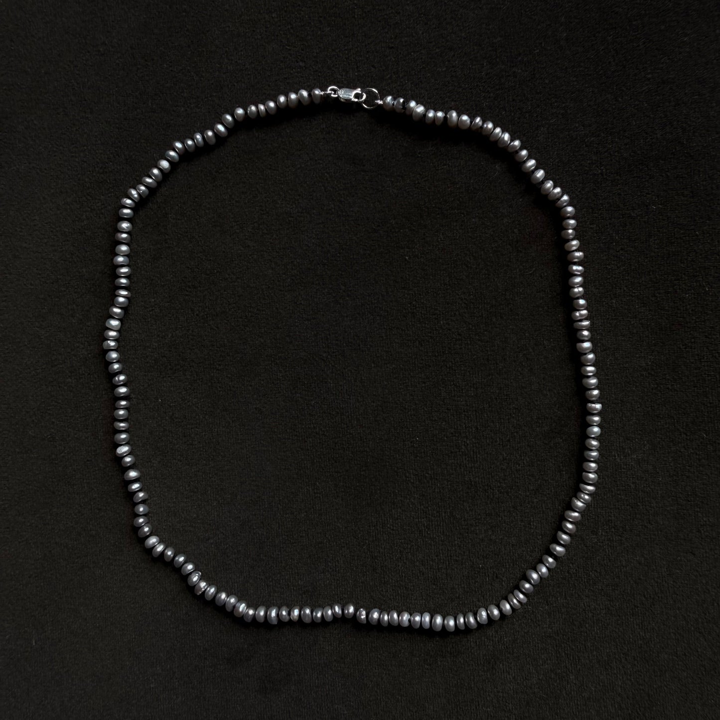 A single-strand beaded necklace made of small, dark gray to black pearls with a silver clasp, displayed on a solid black background.