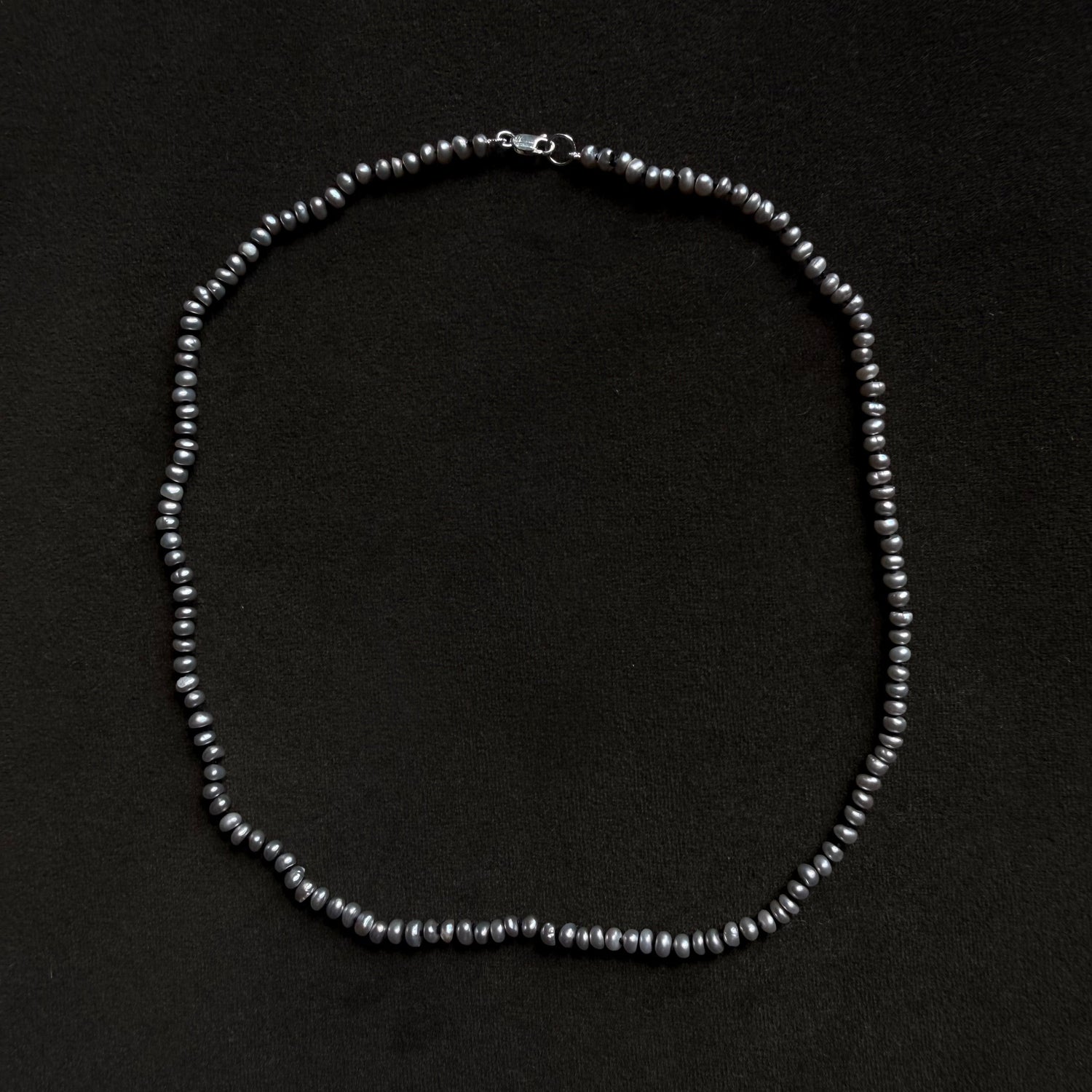 A single-strand beaded necklace made of small, dark gray to black pearls with a silver clasp, displayed on a solid black background.