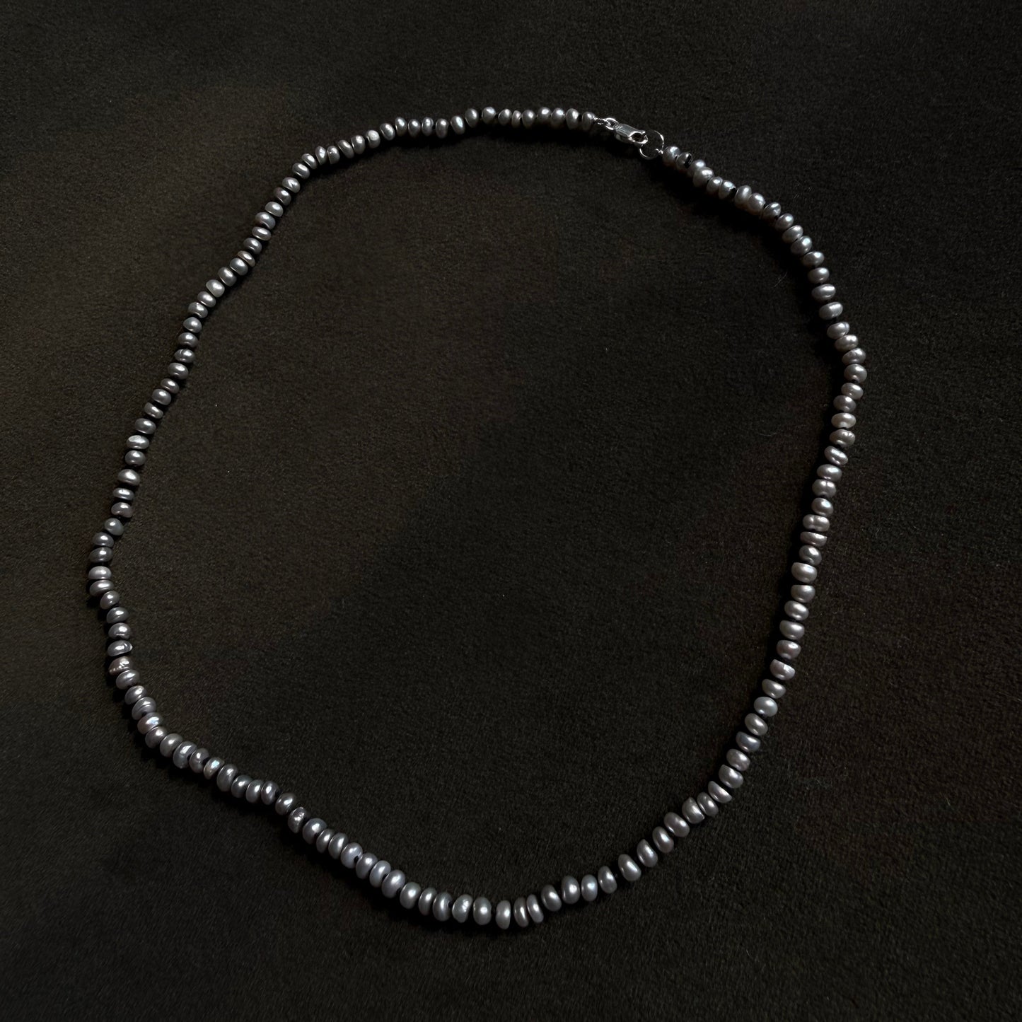 A beaded necklace made of small dark gray to black potato pearls, arranged in an oval shape on a dark textured background.