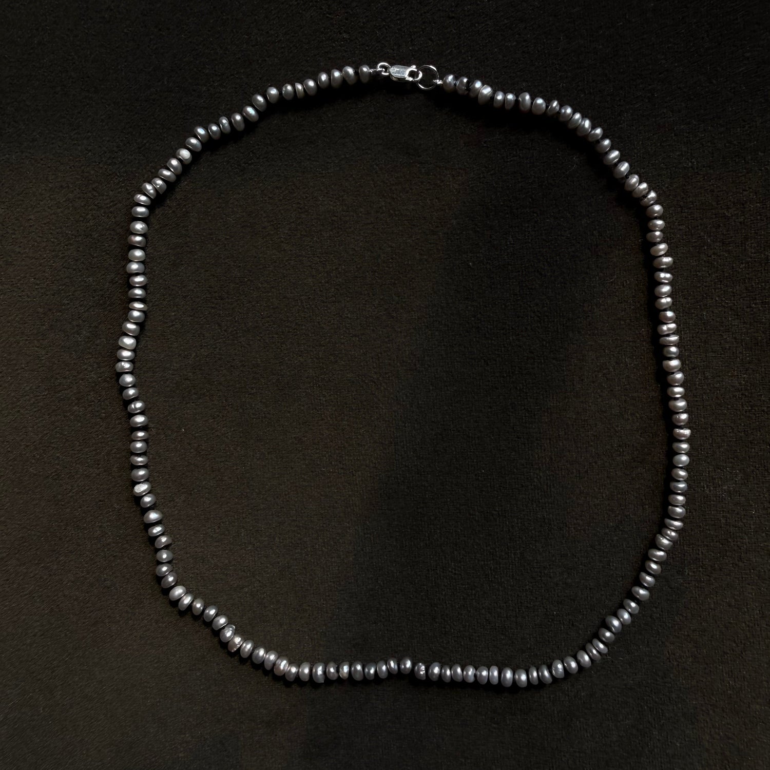 A beaded necklace made of small dark gray metallic beads arranged in an oval shape on a dark textured background.