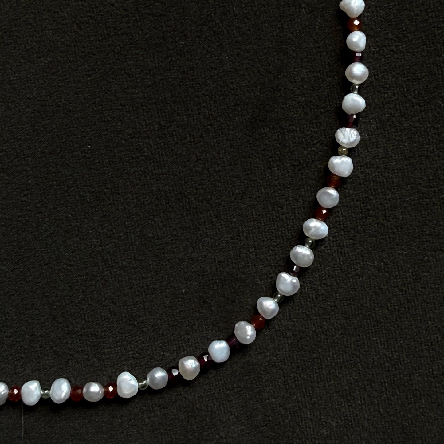 A beaded necklace featuring irregular white baroque pearls separated by small silver spacer beads and dark red faceted beads, displayed on a textured black background.