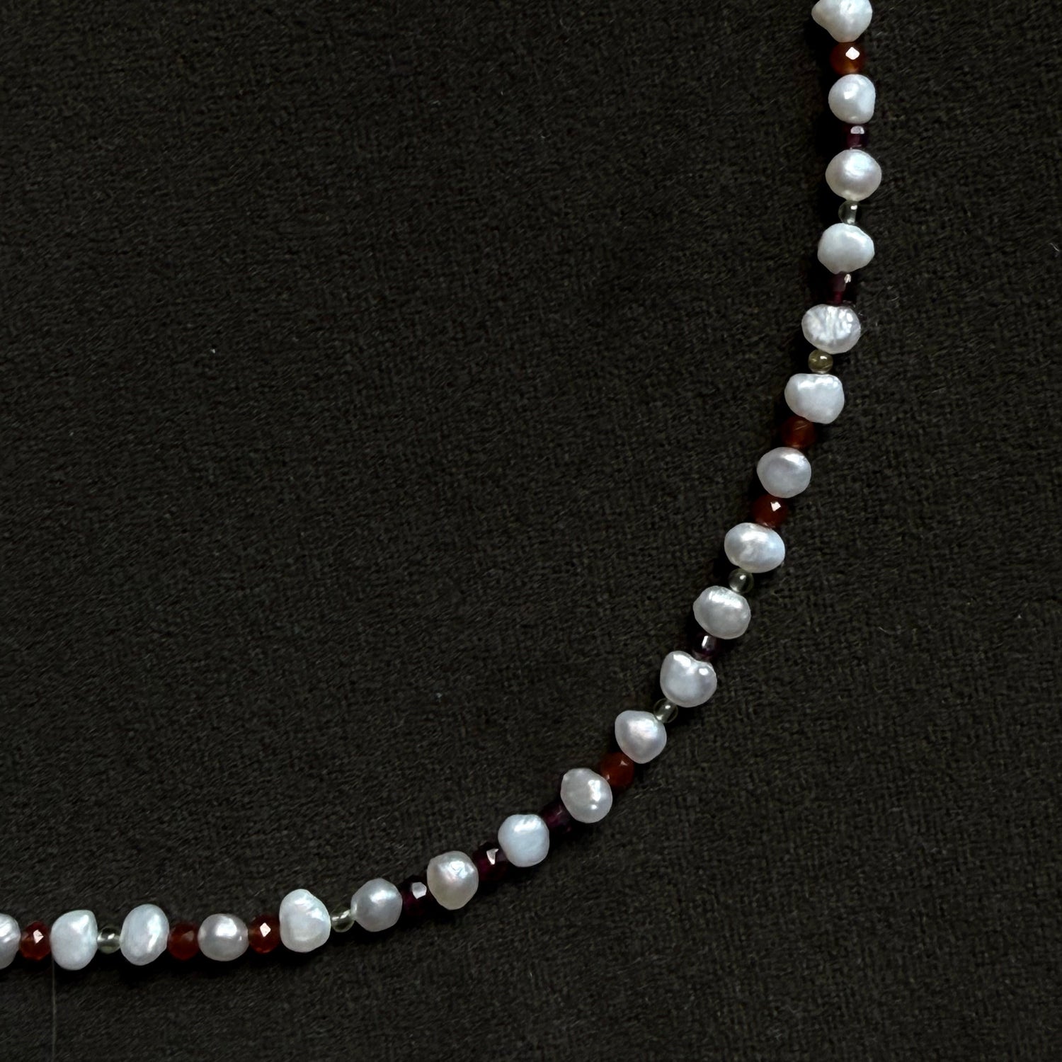 A beaded necklace featuring irregular white baroque pearls separated by small silver spacer beads and dark red faceted beads, displayed on a textured black background.
