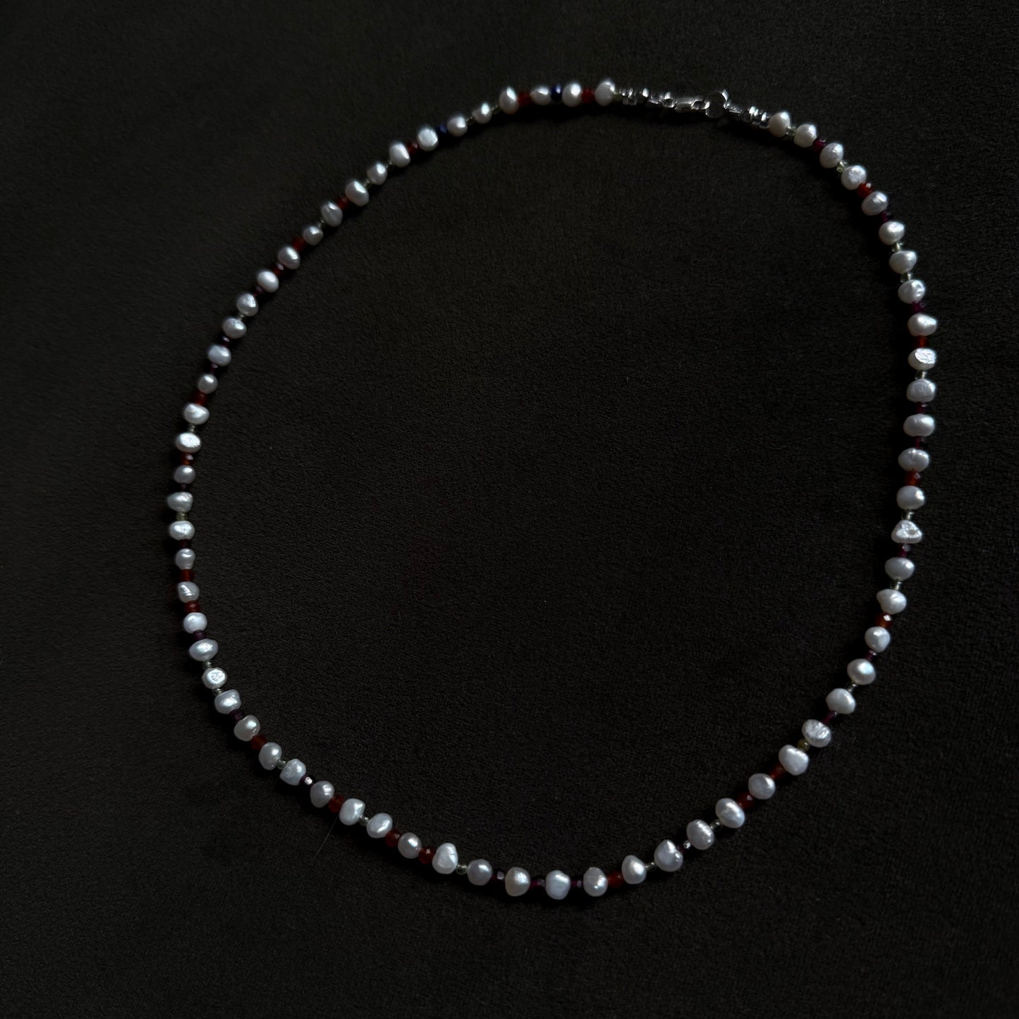 A beaded necklace made of irregular, faceted grayish-white beads with small dark red accents, displayed on a solid black background.