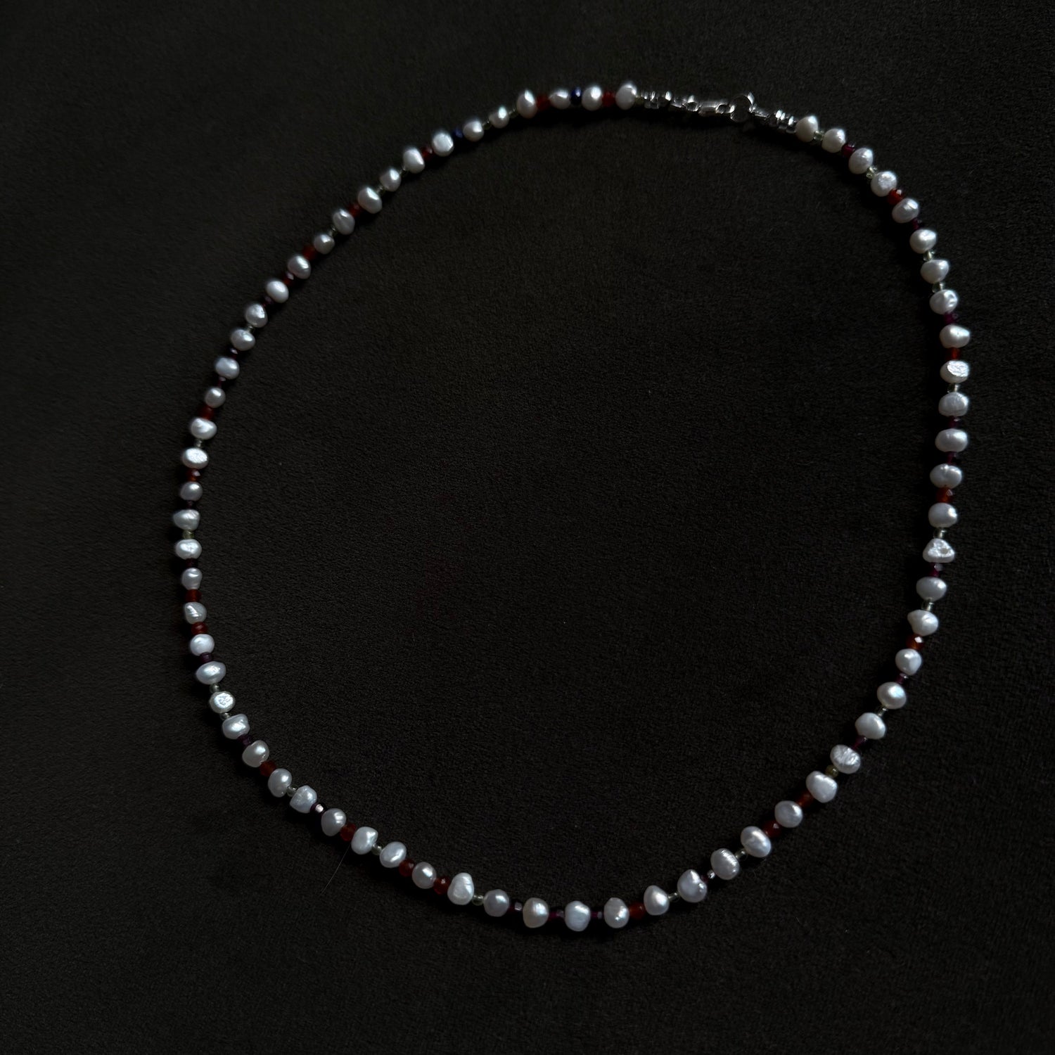 A beaded necklace made of irregular, faceted grayish-white beads with small dark red accents, displayed on a solid black background.