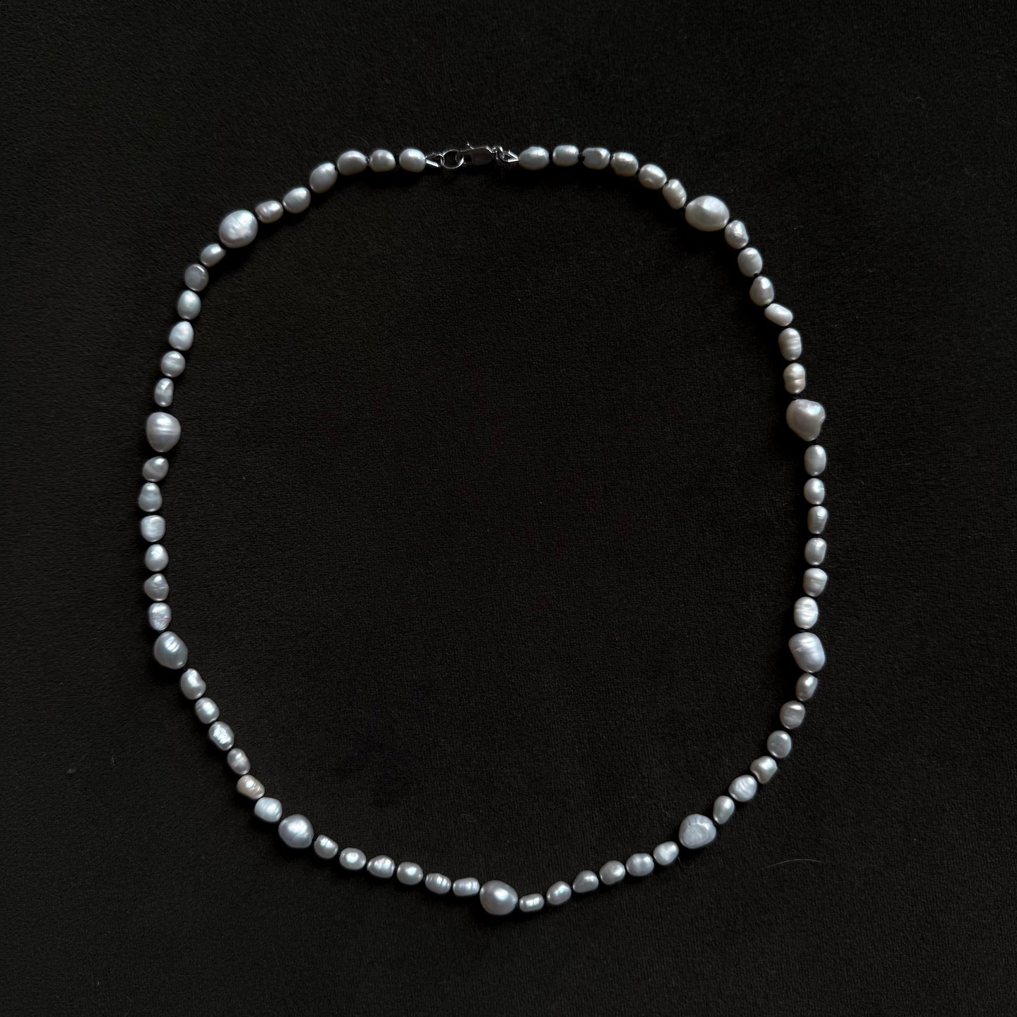 A beaded necklace made of small gray and white baroque pearls arranged in an oval shape on a solid black background.