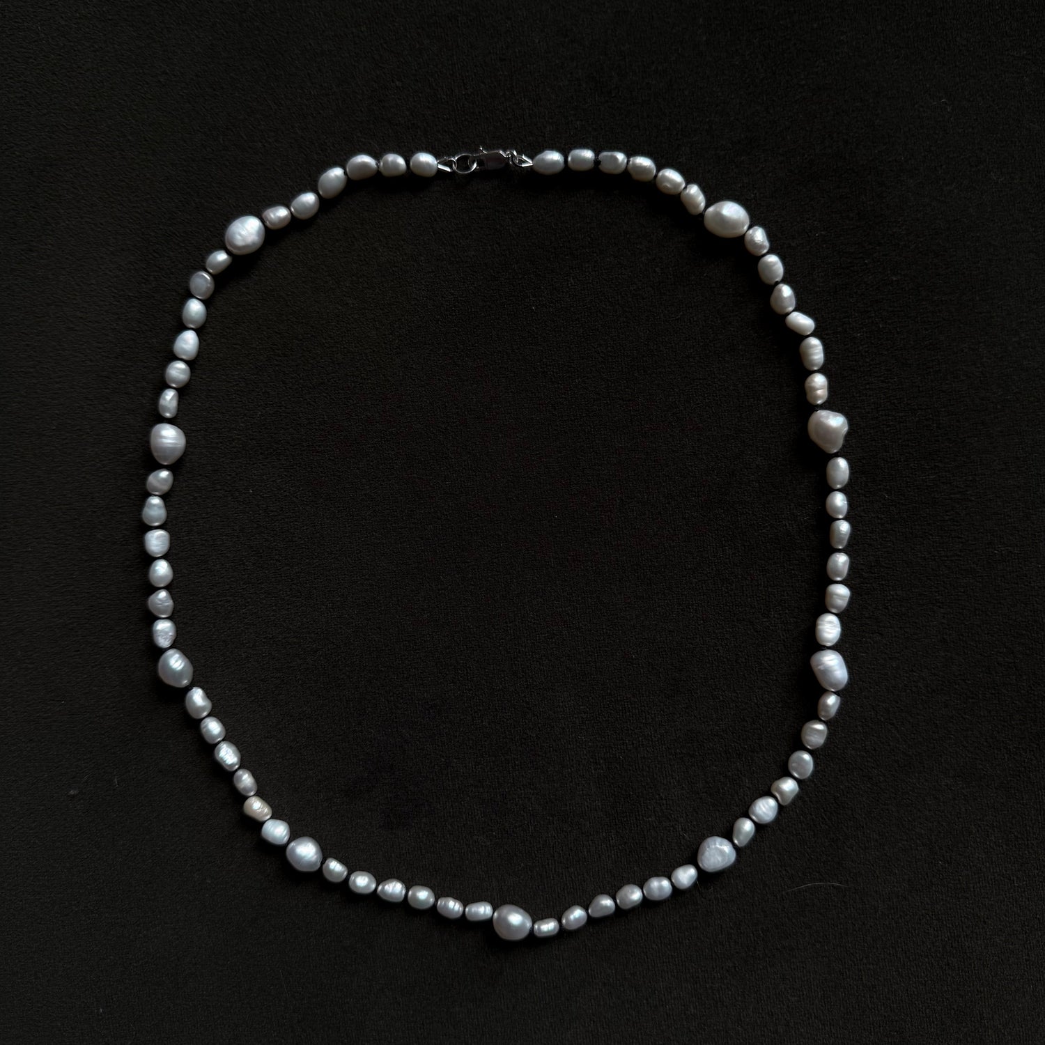 A beaded necklace made of small gray and white baroque pearls arranged in an oval shape on a solid black background.
