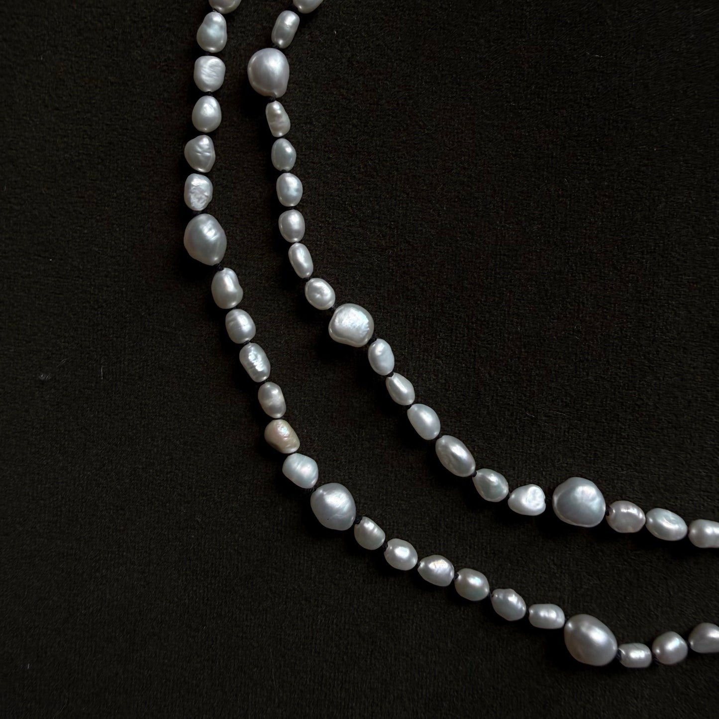 A strand of irregular baroque pearls in a silver-gray luster, laid out in two curved rows on a solid black background.