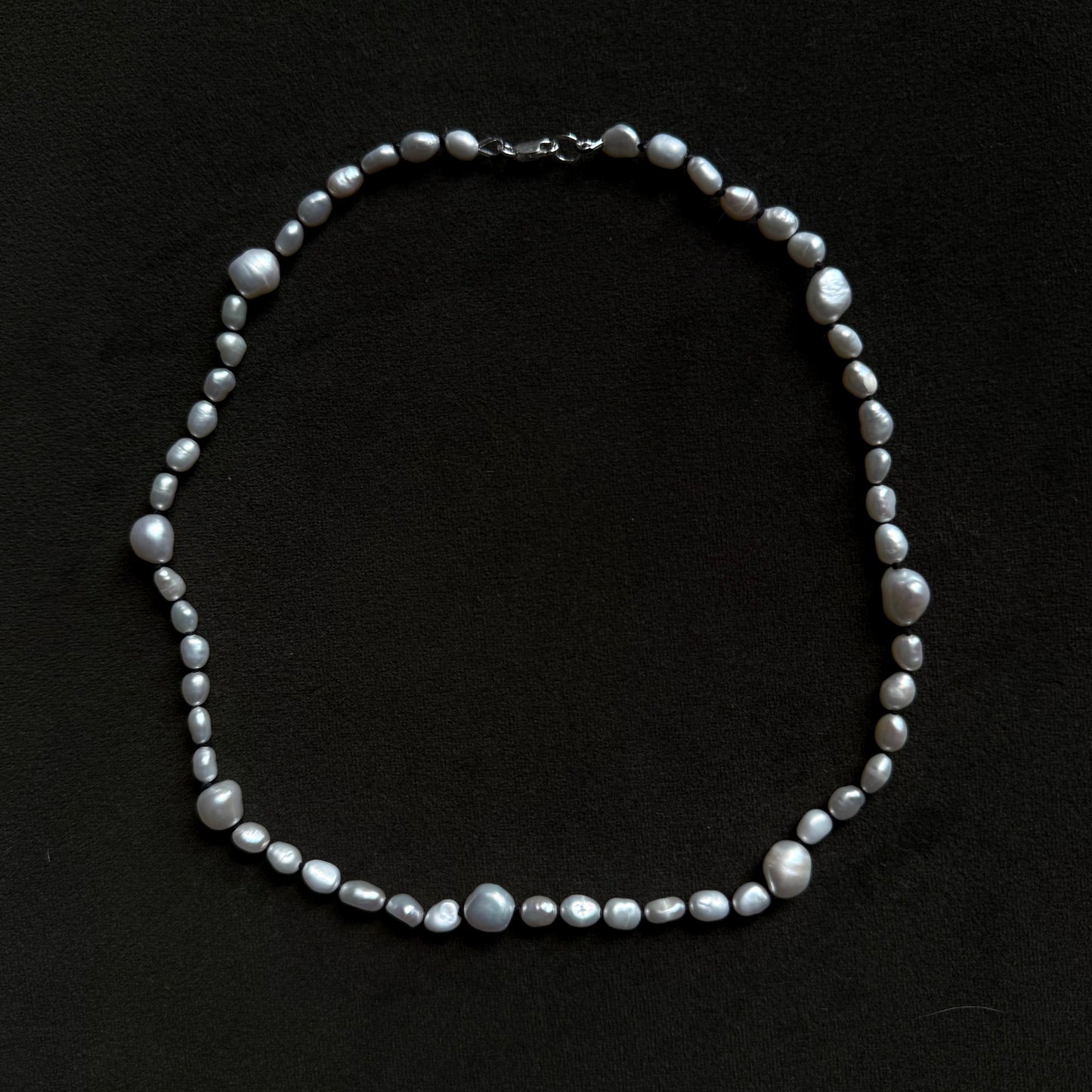 A baroque pearl necklace with irregular, glossy gray-white beads and a silver clasp, displayed on a dark black background.