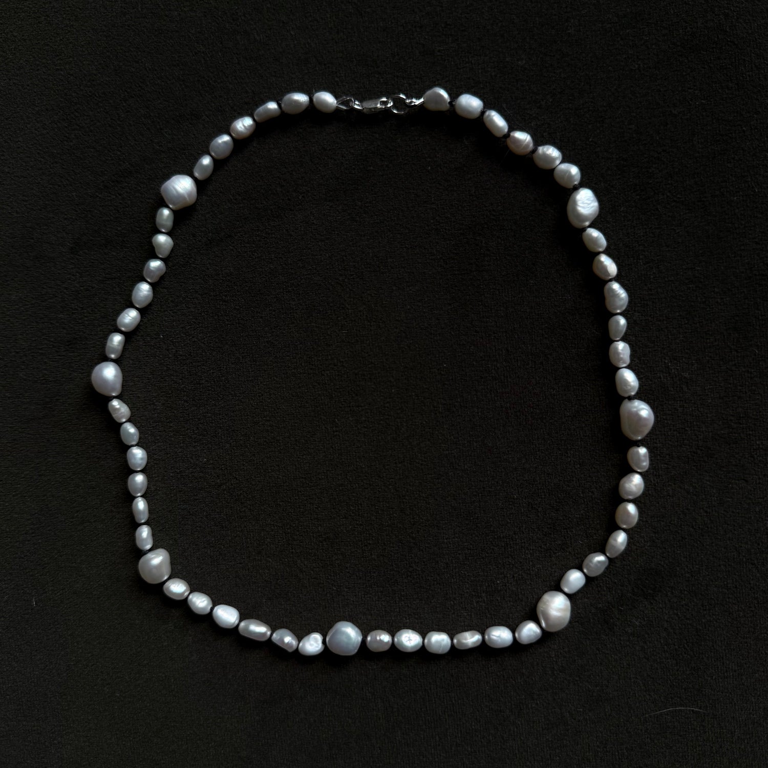 A baroque pearl necklace with irregular, glossy gray-white beads and a silver clasp, displayed on a dark black background.