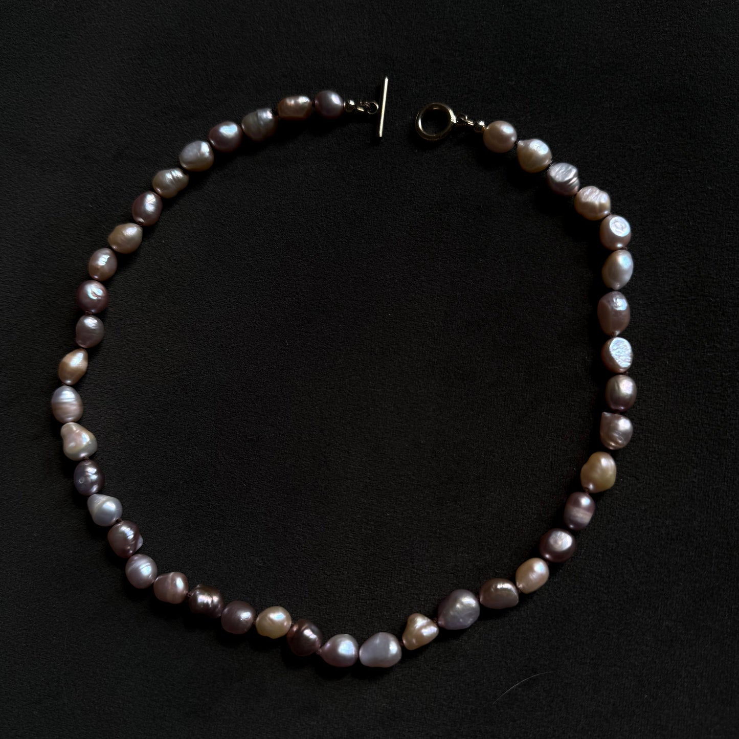 A baroque pearl necklace with irregular, polished beads in shades of gray, purple, and brown, featuring a gold-tone toggle clasp, displayed on a dark black background.