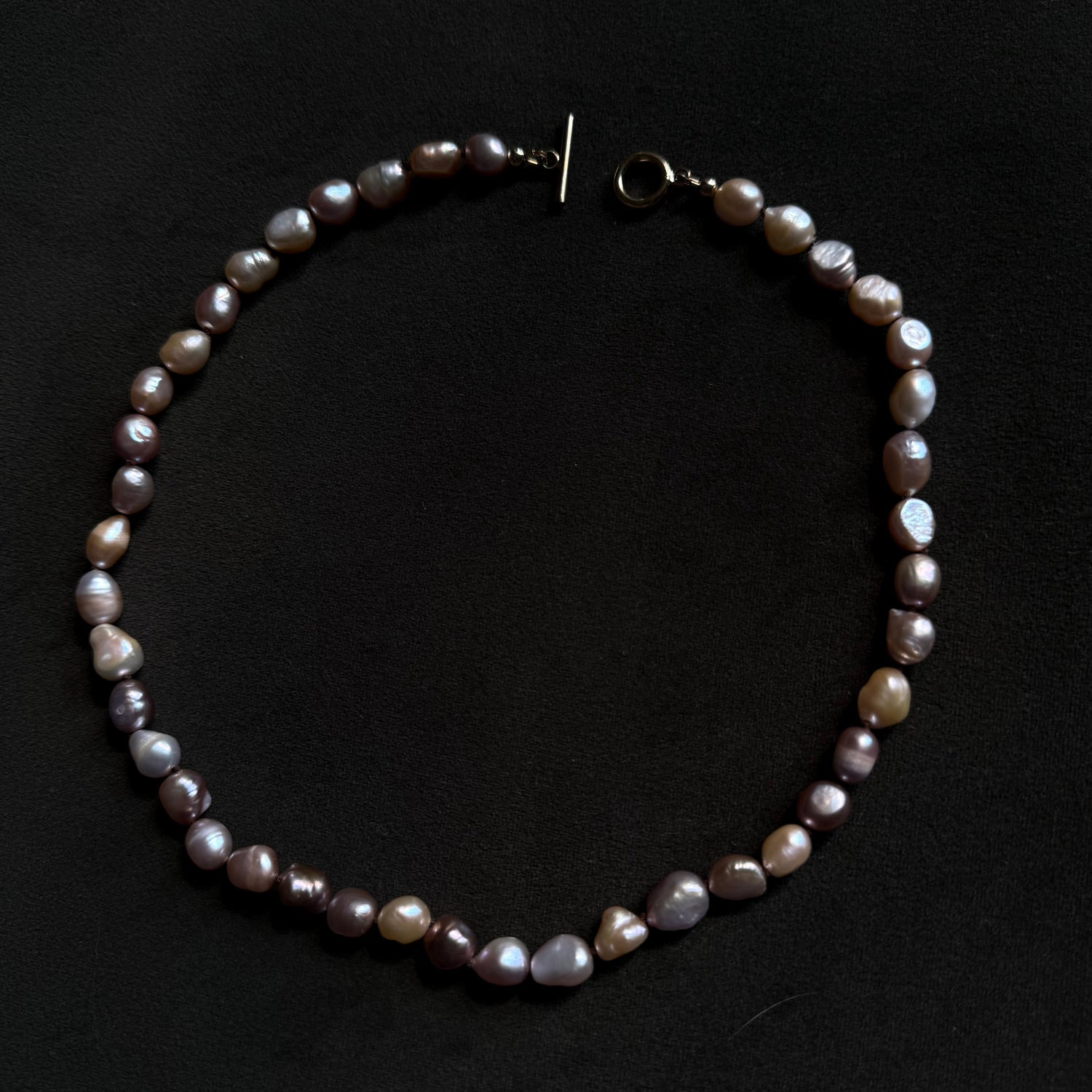 A baroque pearl necklace with irregular, polished beads in shades of gray, purple, and brown, featuring a gold-tone toggle clasp, displayed on a dark black background.