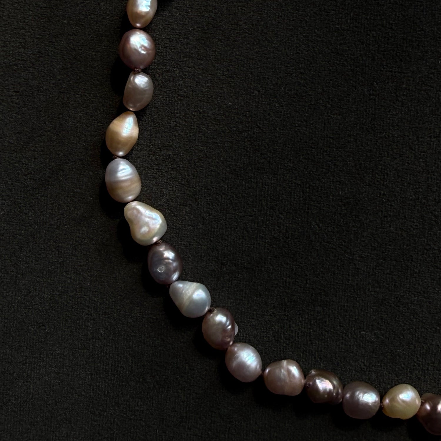 A strand of irregular baroque pearls in muted taupe, lilac, cream, and gray tones, laid out in a curve on a textured black background.