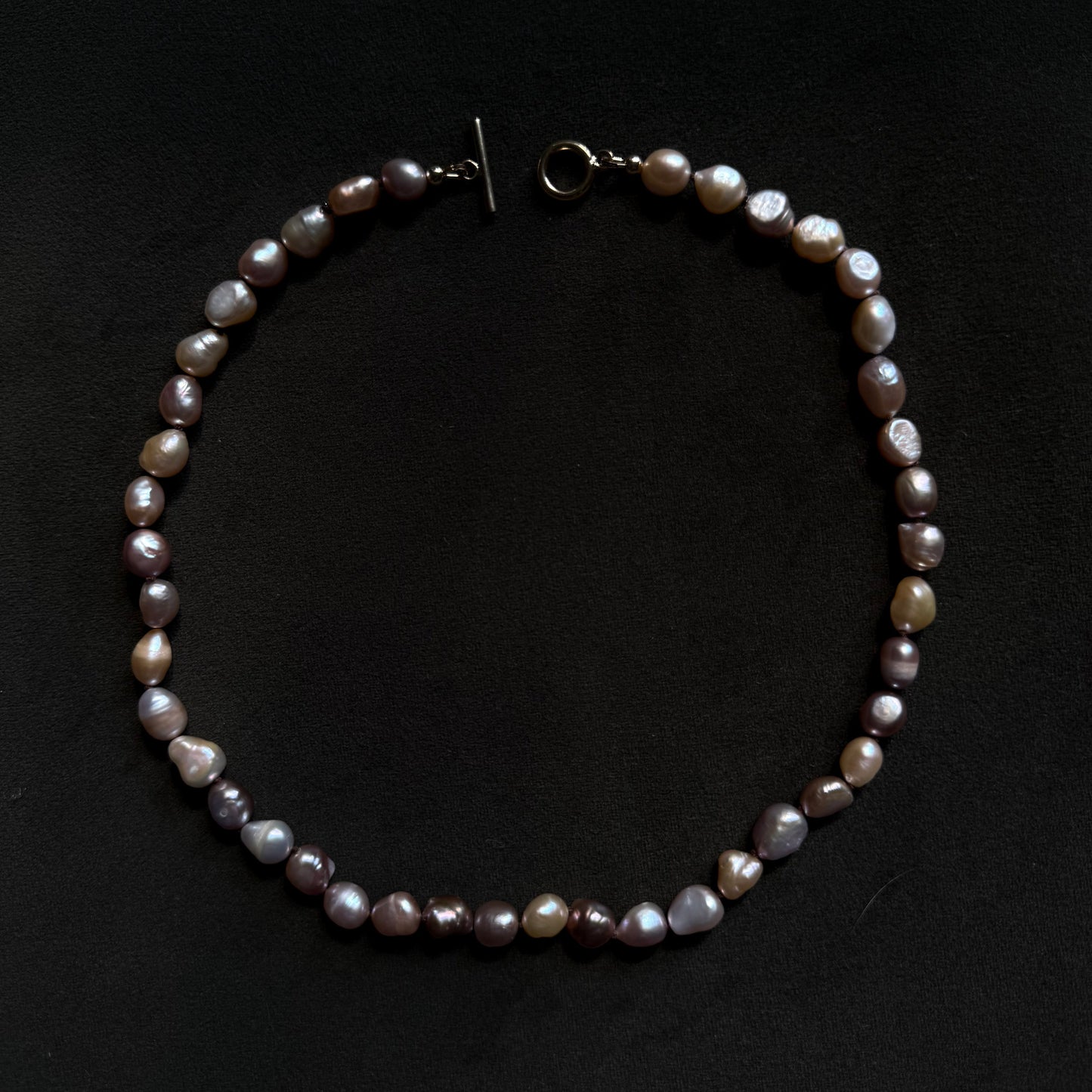 A baroque pearl necklace with irregular, glossy beads in shades of gray, purple, and brown, featuring a dark metal toggle clasp, displayed on a dark black background.