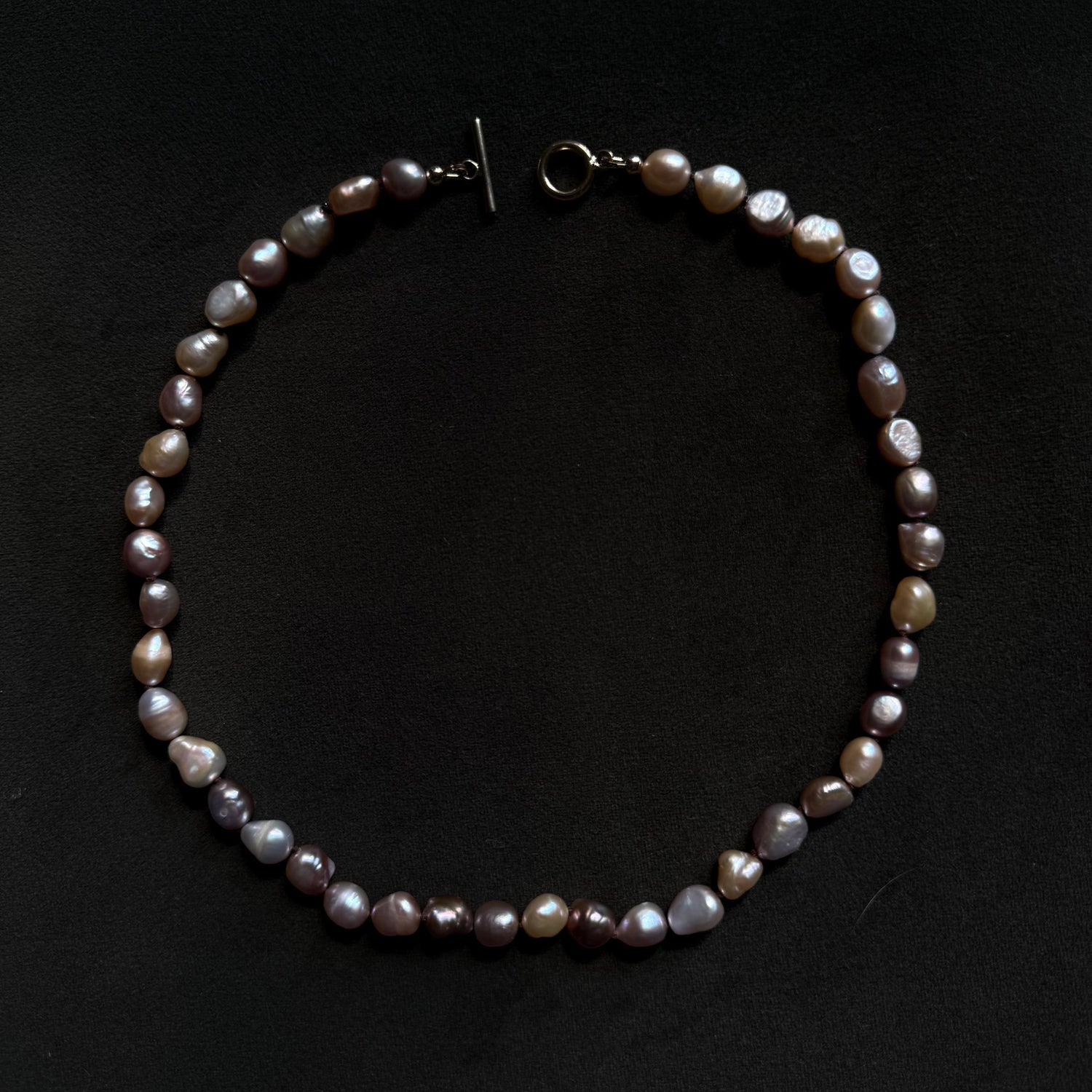 A baroque pearl necklace with irregular, glossy beads in shades of gray, purple, and brown, featuring a dark metal toggle clasp, displayed on a dark black background.
