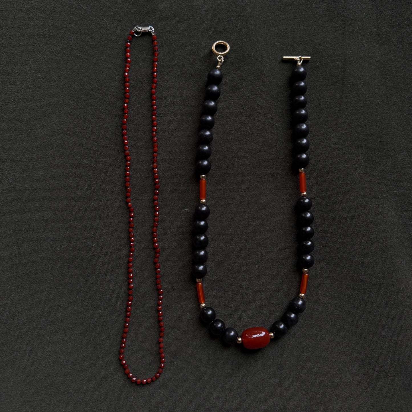Two beaded necklaces laid flat on a dark charcoal background. One necklace is made of small red beads, and the other is made of dark navy or black beads with red spacer sections and a large red oval bead at the bottom.