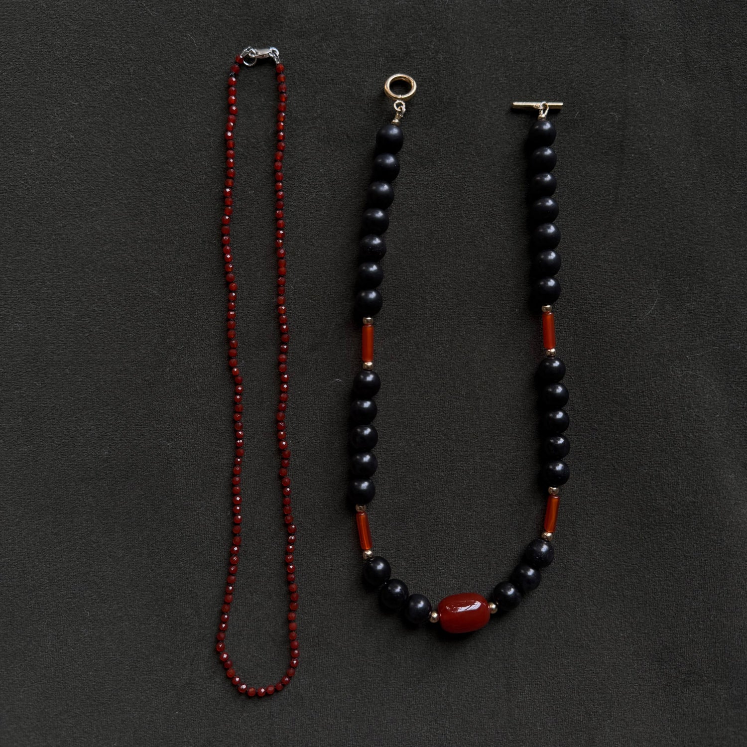 Two beaded necklaces laid flat on a dark charcoal background. One necklace is made of small red beads, and the other is made of dark navy or black beads with red spacer sections and a large red oval bead at the bottom.