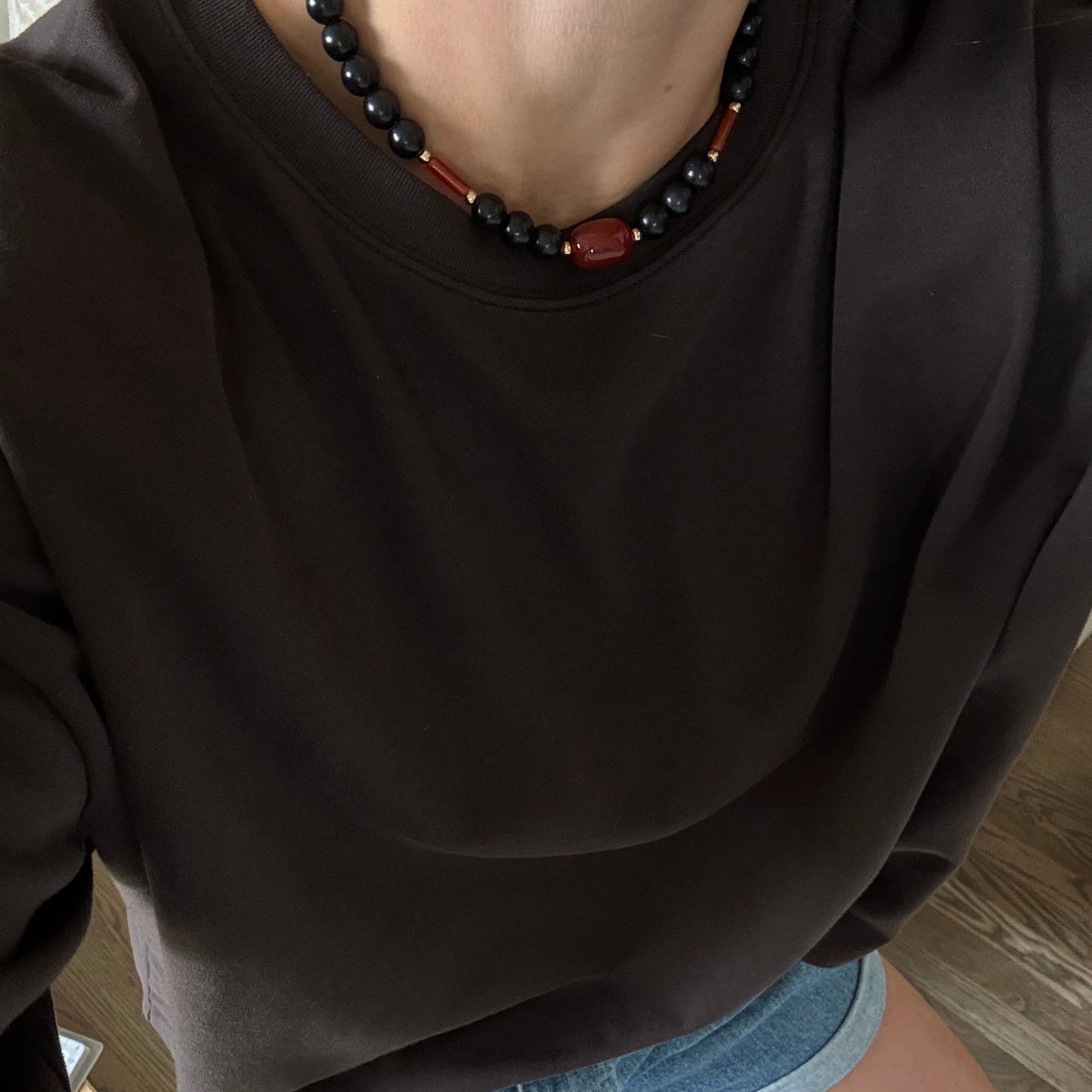 A person wearing a dark brown crewneck sweatshirt and denim shorts, shown from a top-down perspective while kneeling on a wooden floor. They are also wearing a black beaded necklace with a red accent.