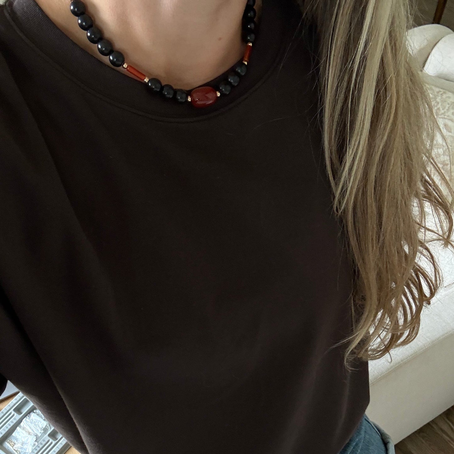 A person wearing an oversized dark brown crewneck t-shirt and denim shorts, accessorized with a black and red beaded necklace, standing indoors on a wooden floor.