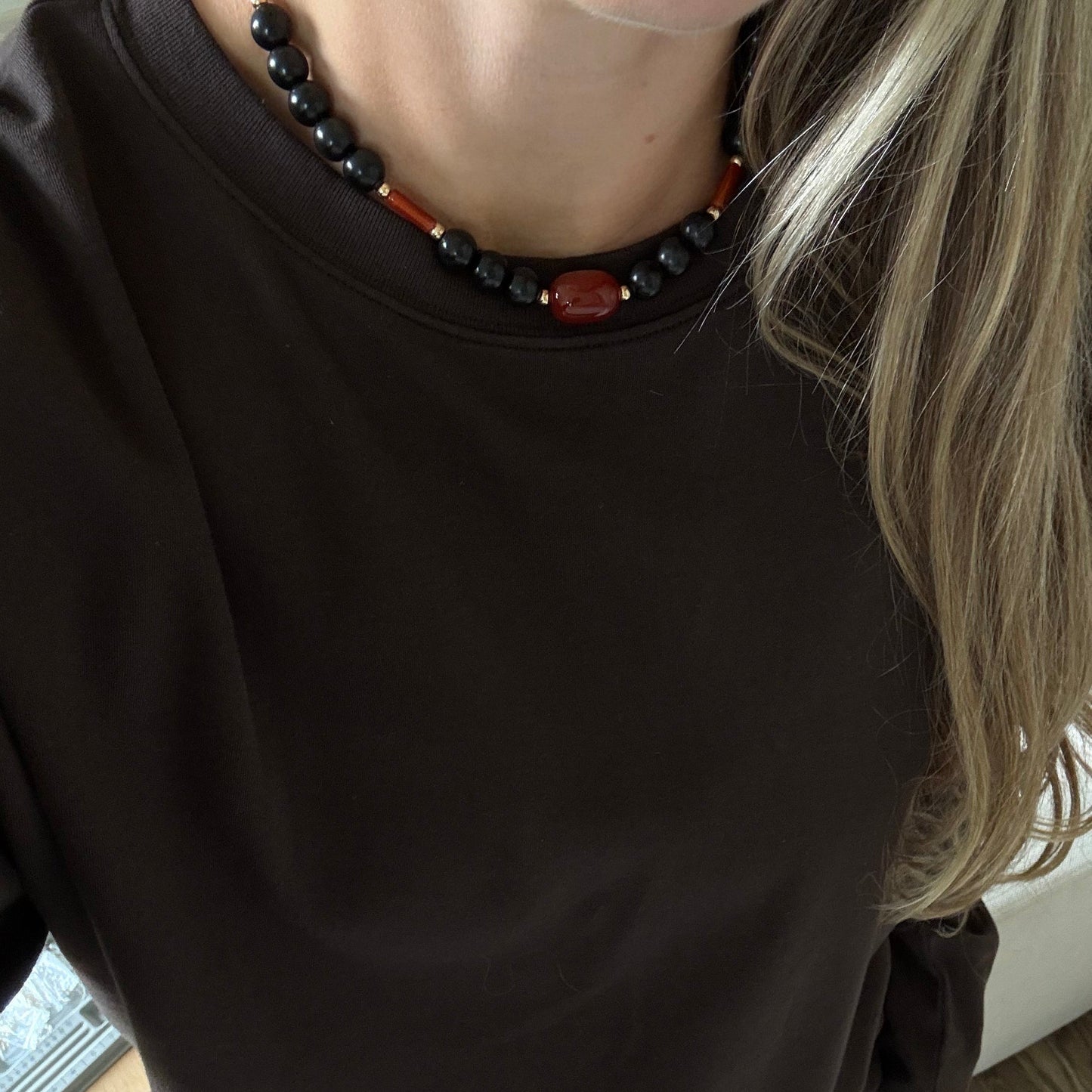A person wearing a dark brown long-sleeve crewneck top, styled with a black and red beaded necklace and denim shorts, standing on a wooden floor.
