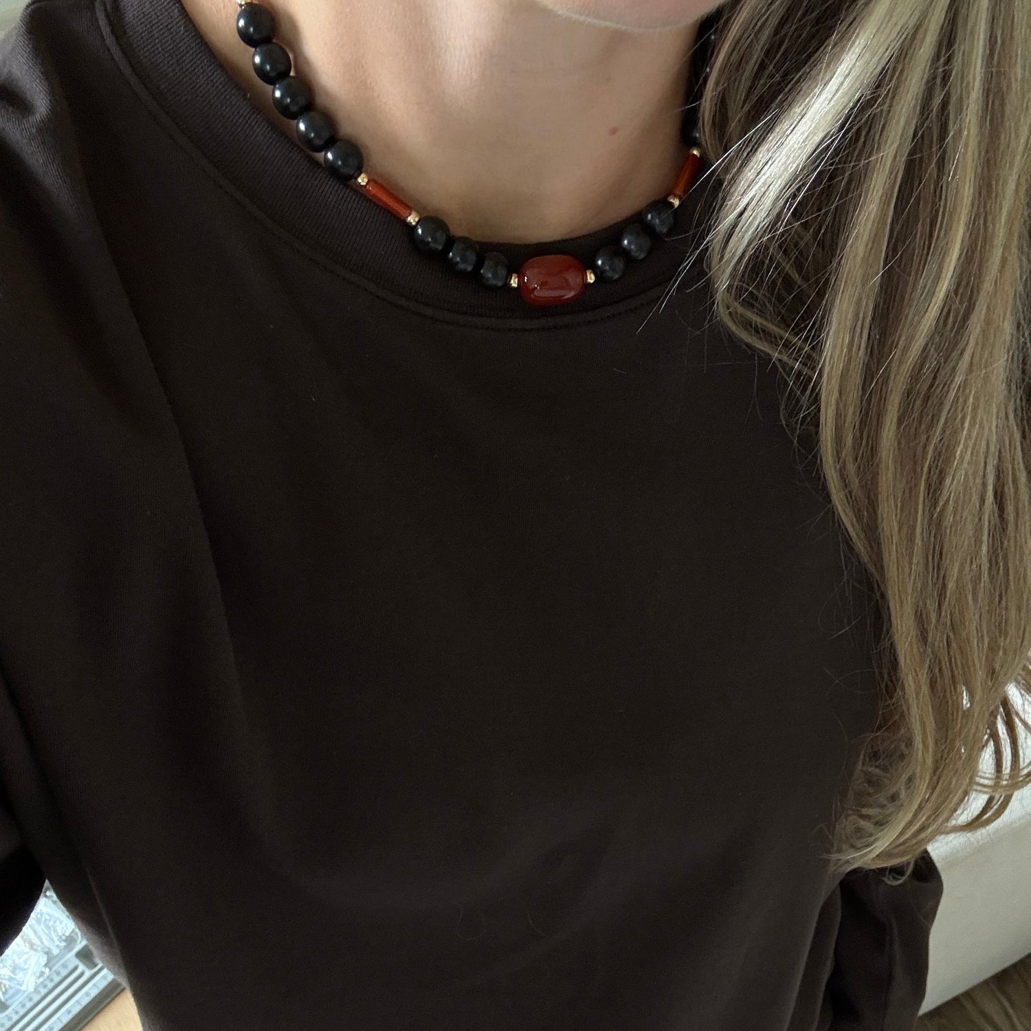 A person wearing a dark brown long-sleeve crewneck top, styled with a black and red beaded necklace and denim shorts, standing on a wooden floor.