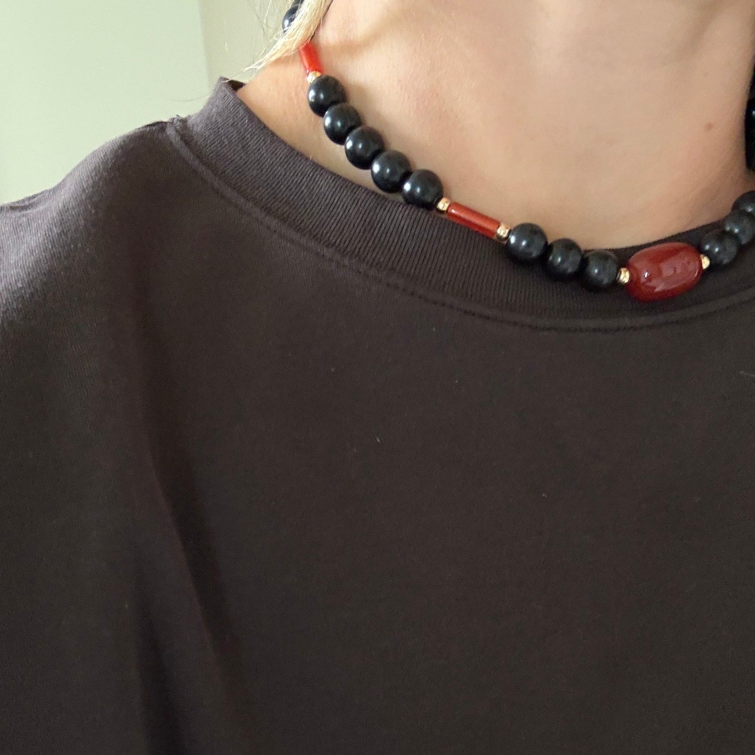 A close-up of a person wearing a dark brown crewneck sweatshirt and a beaded necklace with black round beads and red accents.