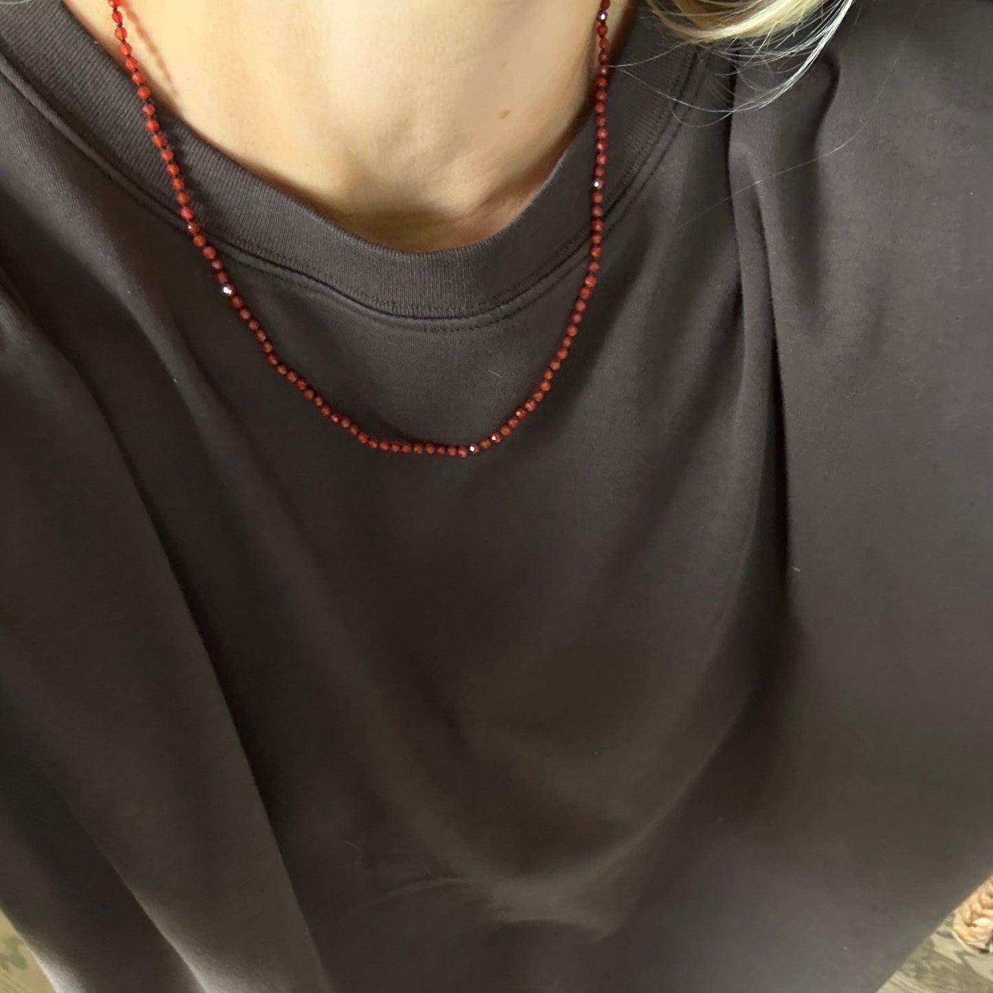 A close-up of a person wearing an oversized dark brown crewneck t-shirt and a red beaded necklace, standing on a wooden floor.