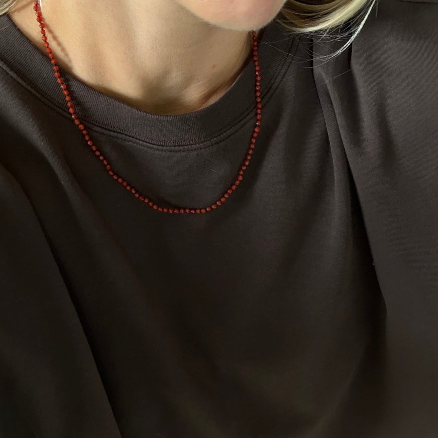A close-up of a person wearing a dark brown crewneck sweatshirt and a red beaded necklace.