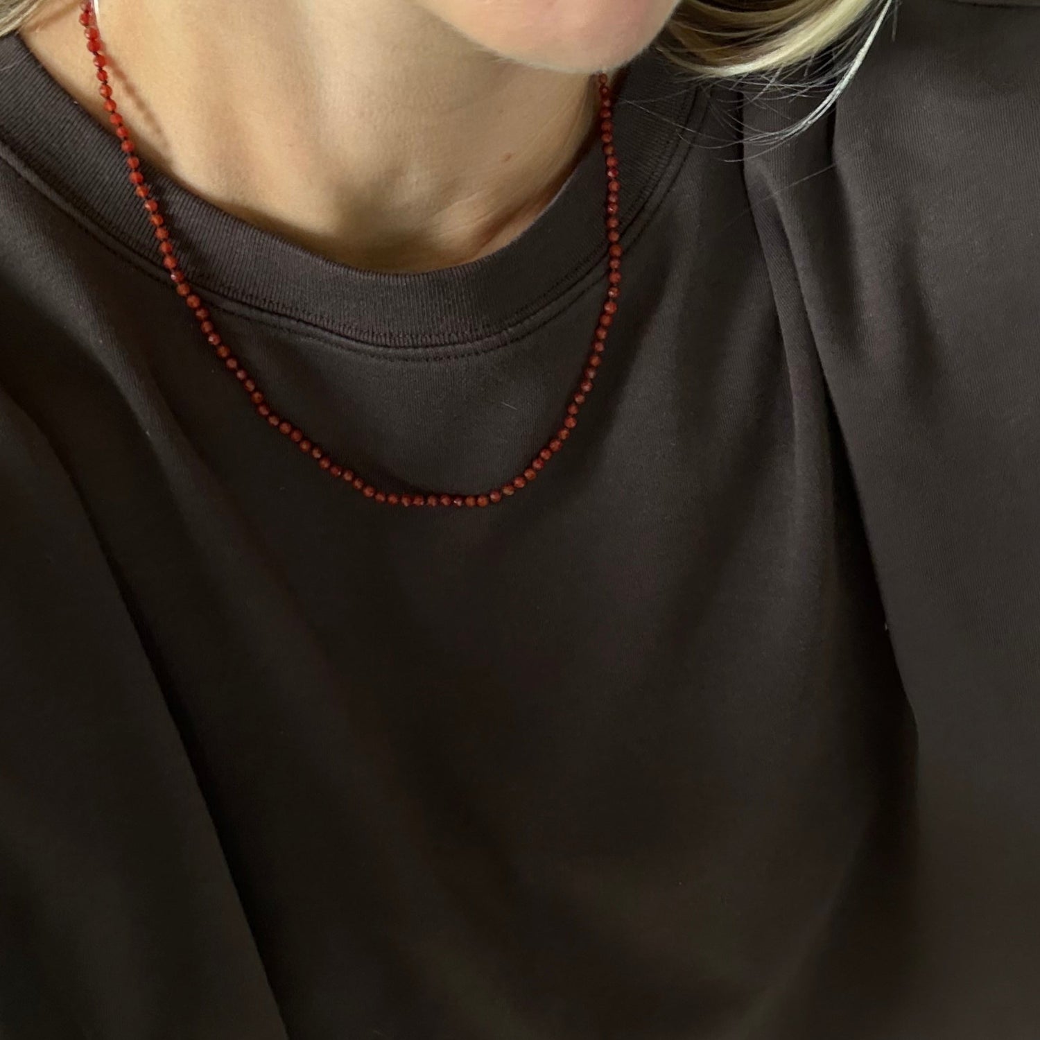 A close-up of a person wearing a dark brown crewneck sweatshirt and a red beaded necklace.