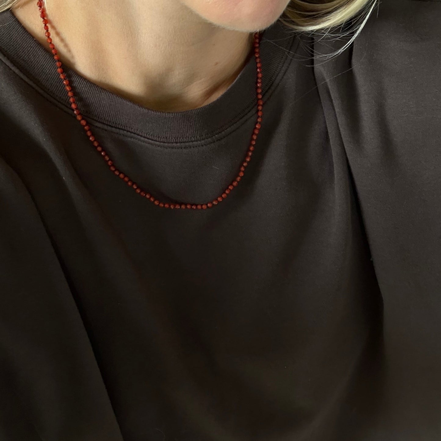 A close-up of a person wearing a dark brown crewneck sweatshirt and a thin red beaded necklace.