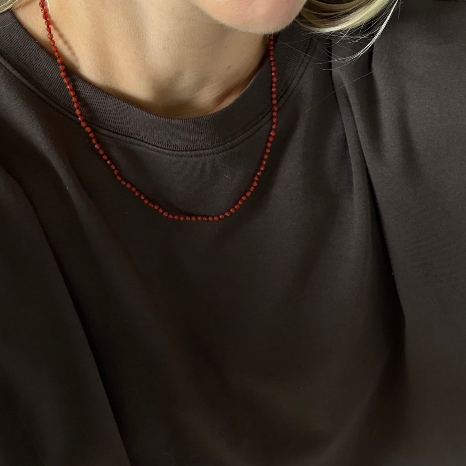 A close-up of a person wearing a dark brown crewneck sweatshirt and a thin red beaded necklace.
