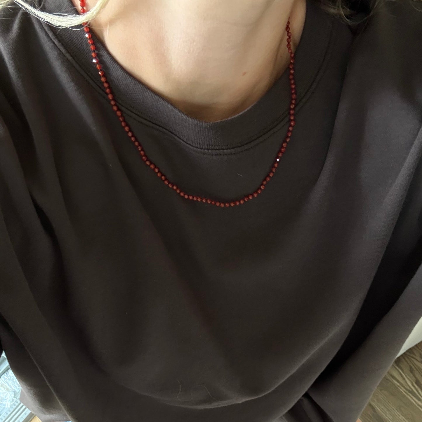 A person wearing an oversized dark brown crewneck t-shirt and a red beaded necklace, standing barefoot on a wood floor.