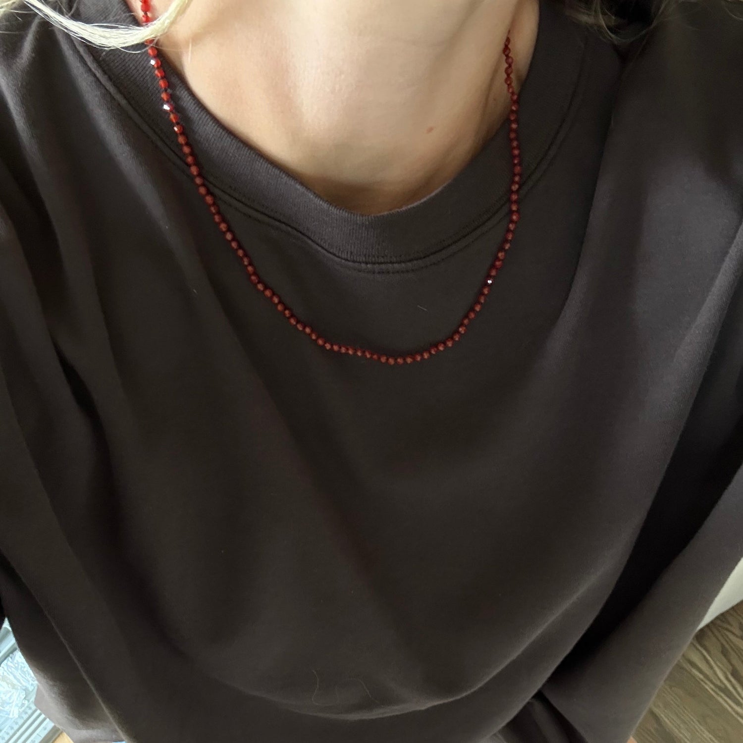 A person wearing an oversized dark brown crewneck t-shirt and a red beaded necklace, standing barefoot on a wood floor.