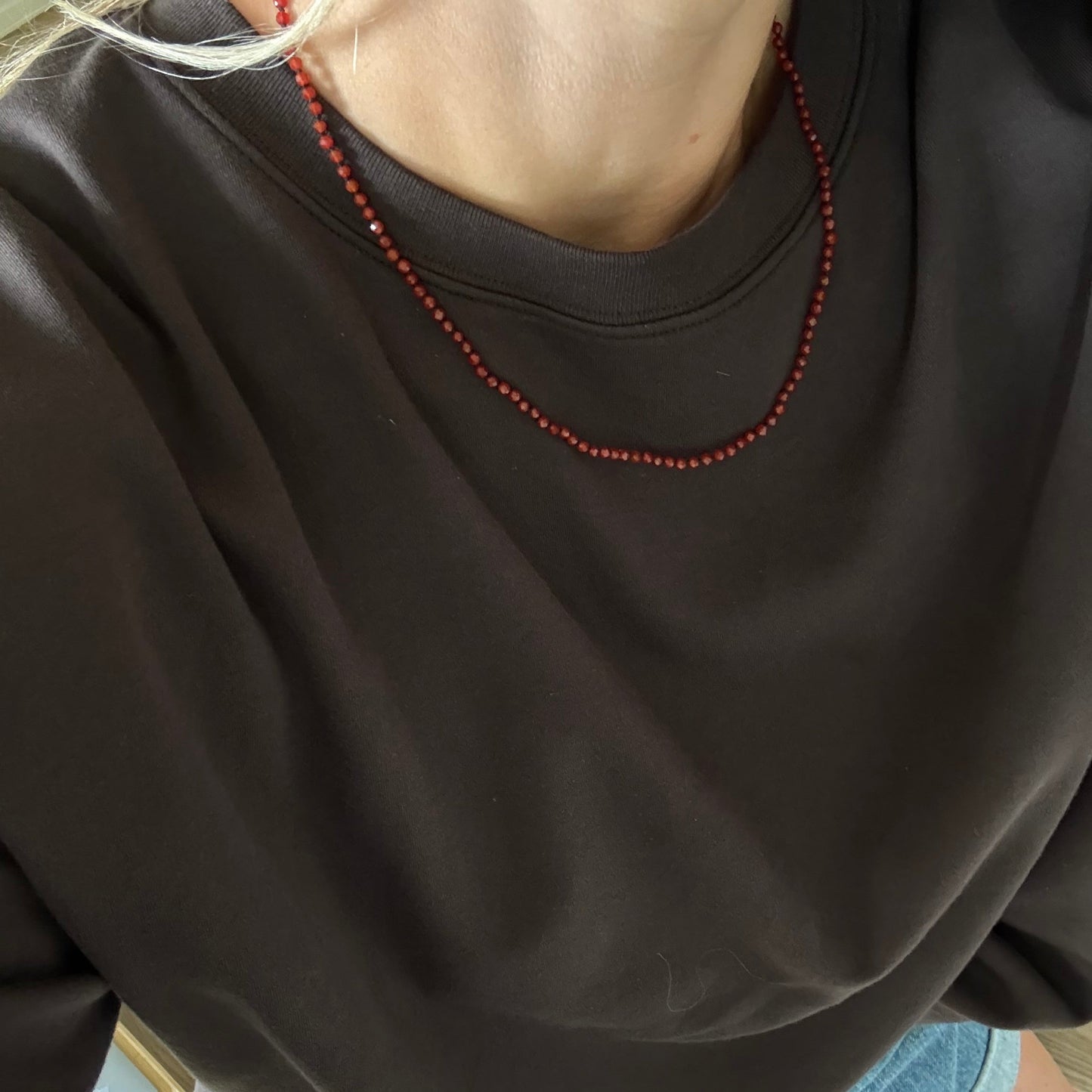 A close-up lifestyle shot of a person wearing a dark brown crewneck sweatshirt and a red beaded necklace.