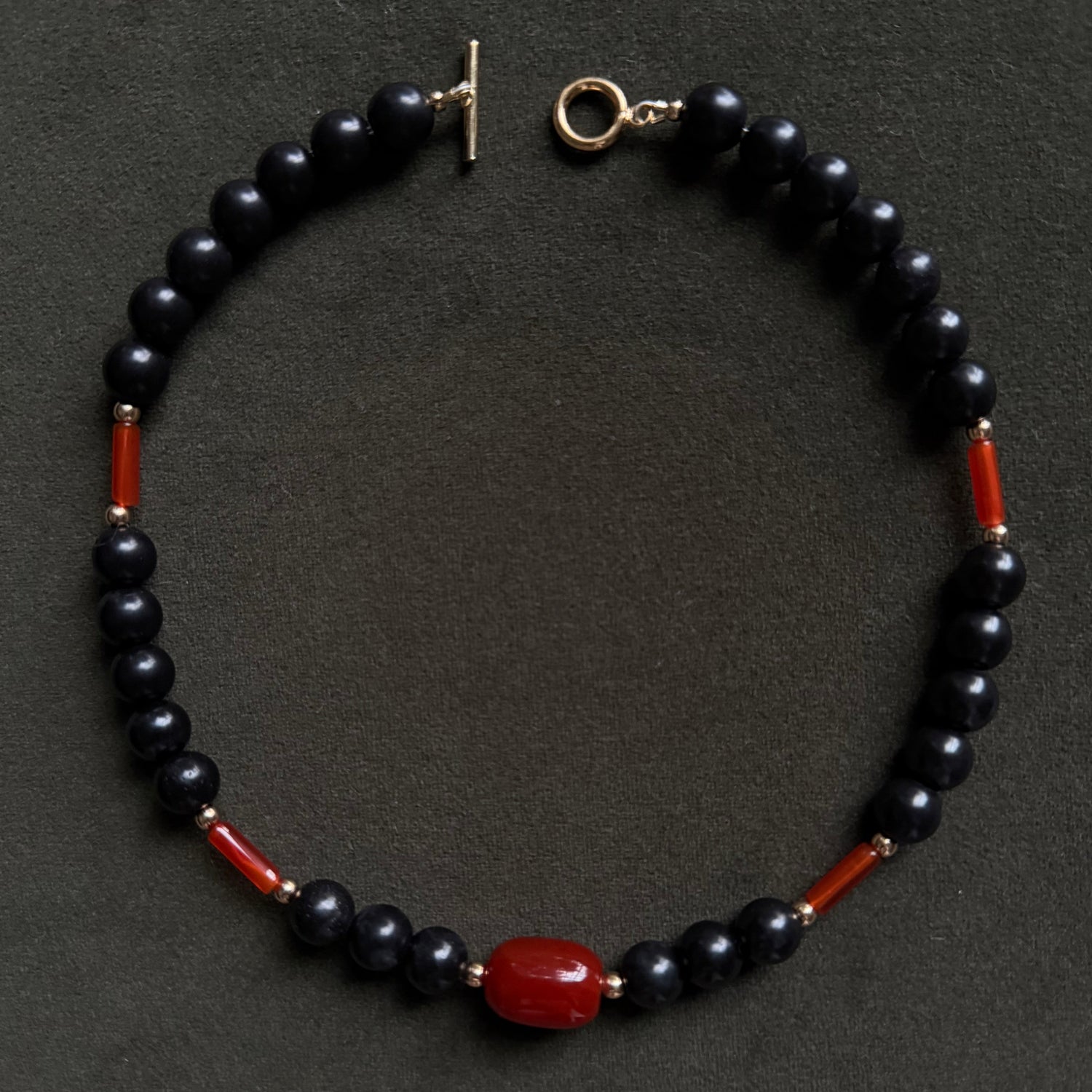 A beaded necklace featuring glossy black round beads, small red cylindrical accents, and a prominent polished red oval centerpiece, displayed on a dark textured background.