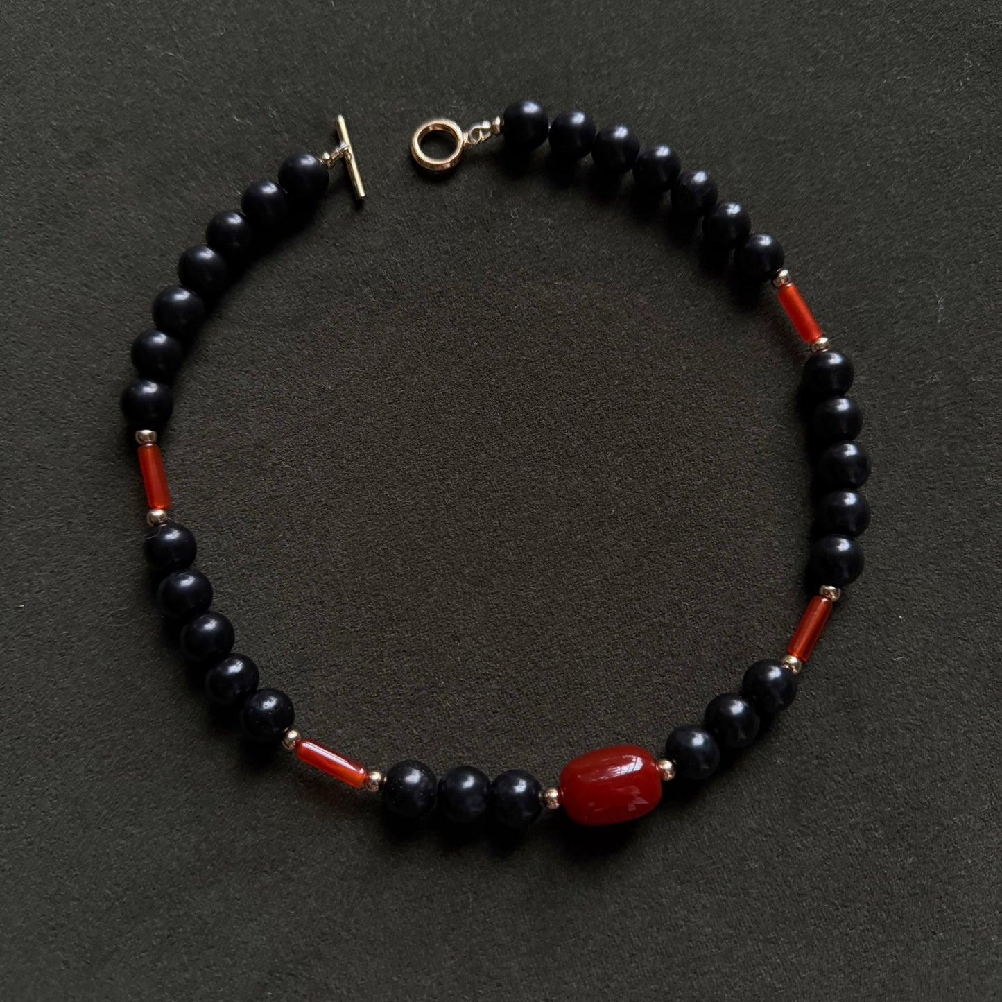 A beaded bracelet made of glossy black round beads with small red spacer beads and a large polished red oval bead at the bottom, featuring a gold-tone toggle clasp, displayed on a dark textured background.