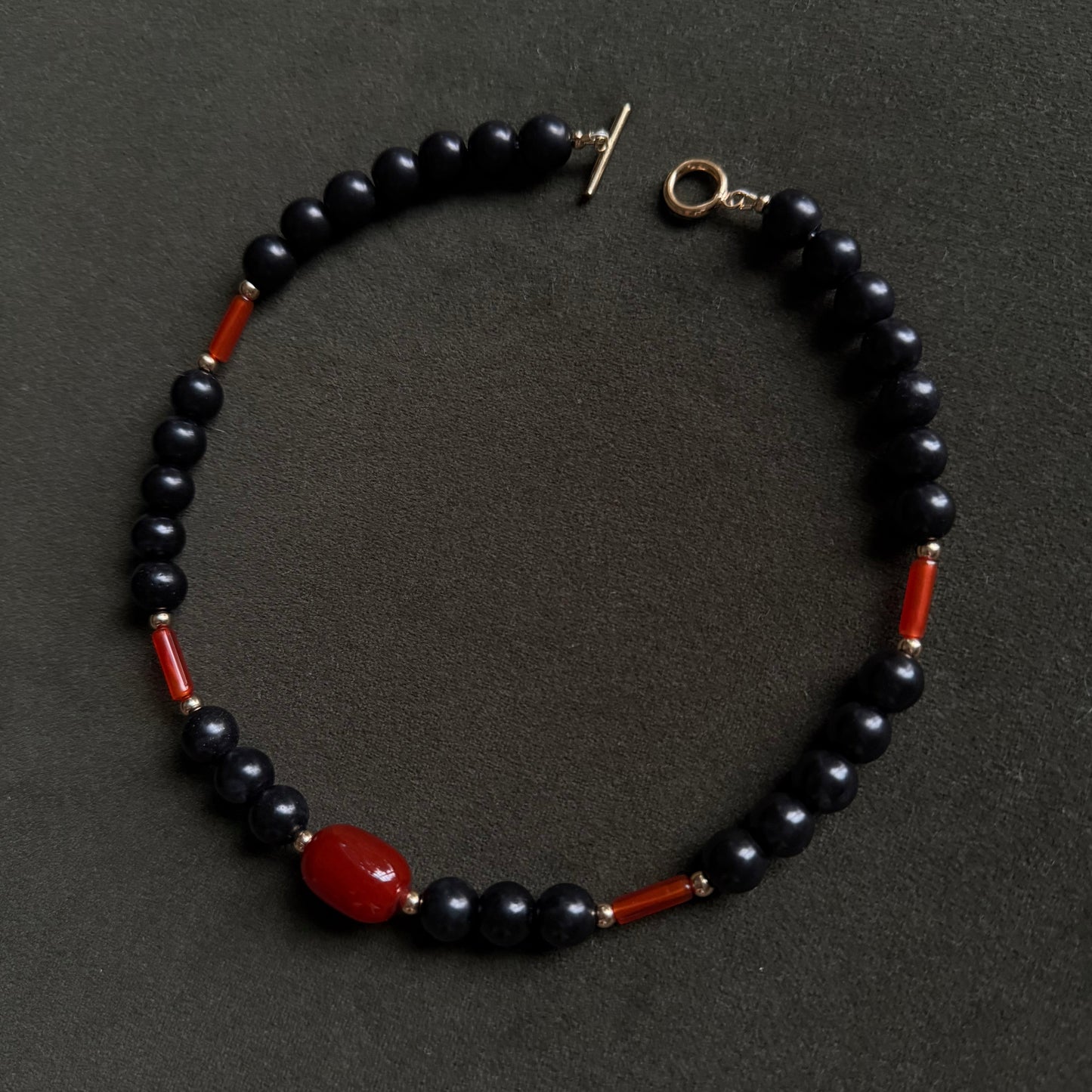 A beaded necklace featuring glossy black round beads, small red cylindrical beads, and a larger polished red oval bead, with a gold-tone toggle clasp, displayed on a dark textured background.
