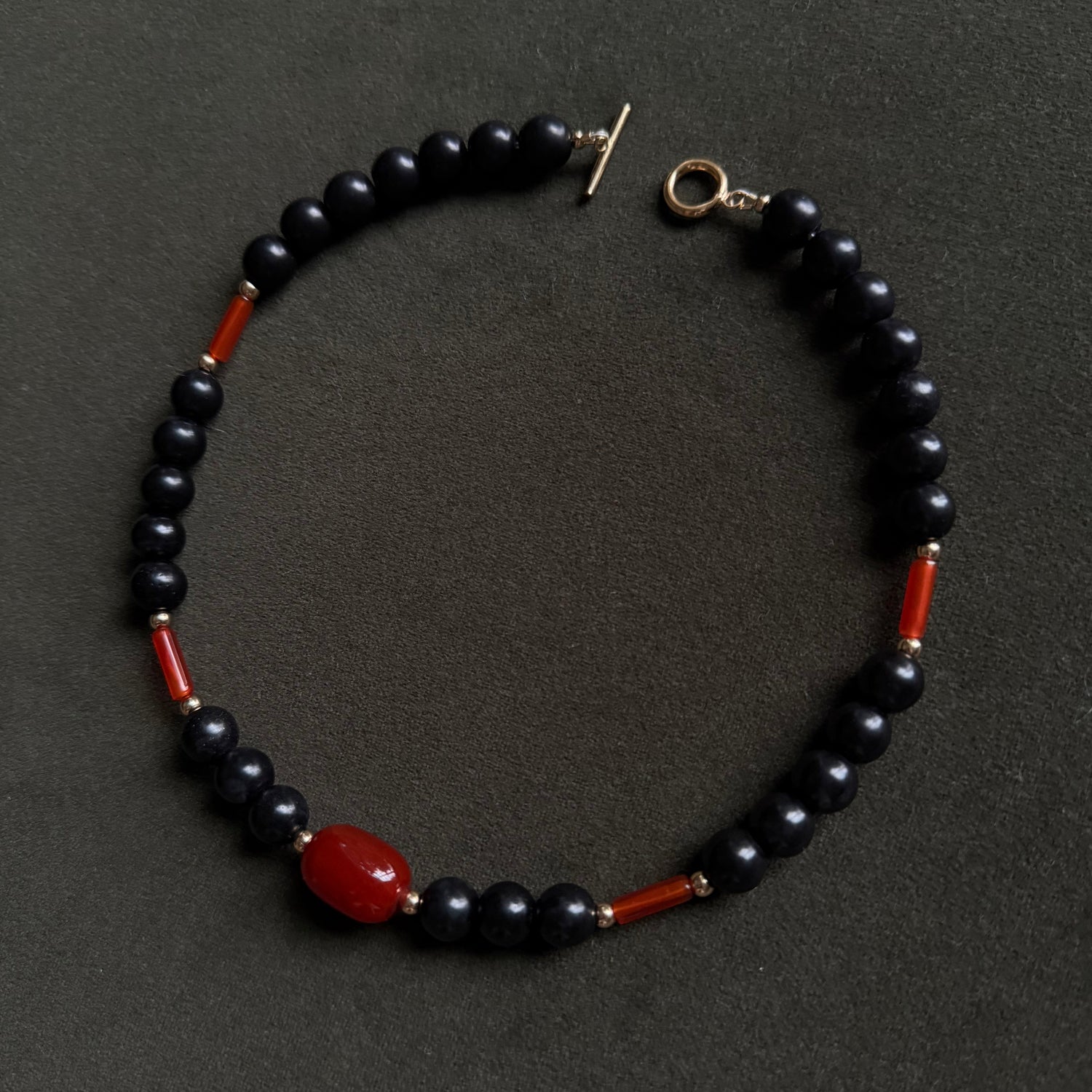A beaded necklace featuring glossy black round beads, small red cylindrical beads, and a larger polished red oval bead, with a gold-tone toggle clasp, displayed on a dark textured background.