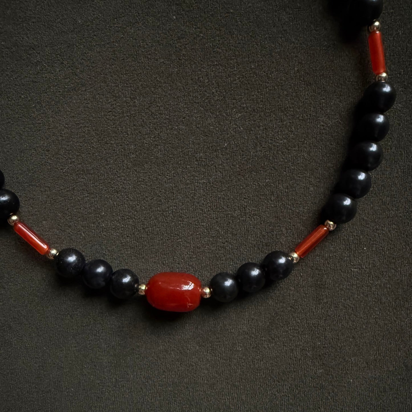 A beaded necklace featuring glossy black round beads, polished red cylindrical spacer beads, and a central red oval bead, displayed on a dark textured background.