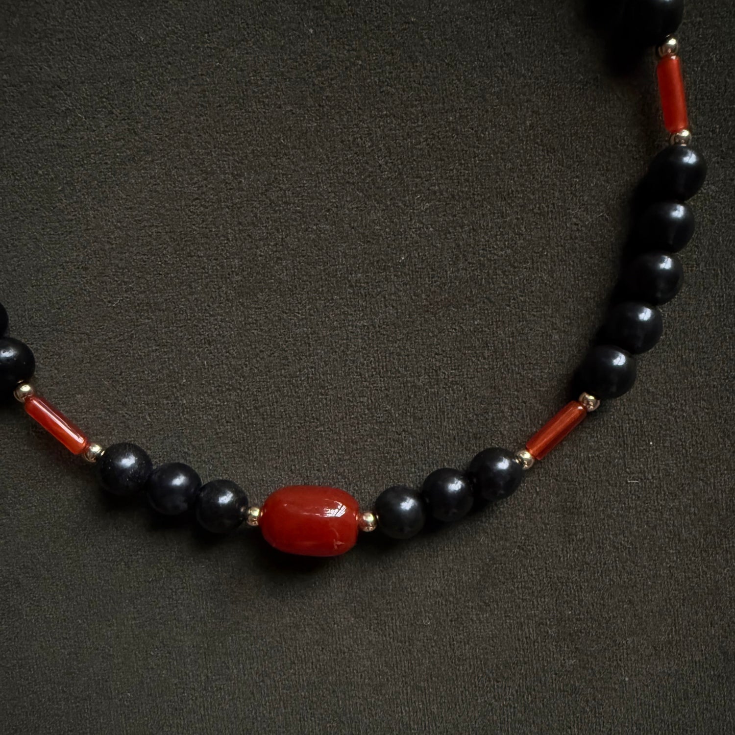 A beaded necklace featuring glossy black round beads, polished red cylindrical spacer beads, and a central red oval bead, displayed on a dark textured background.