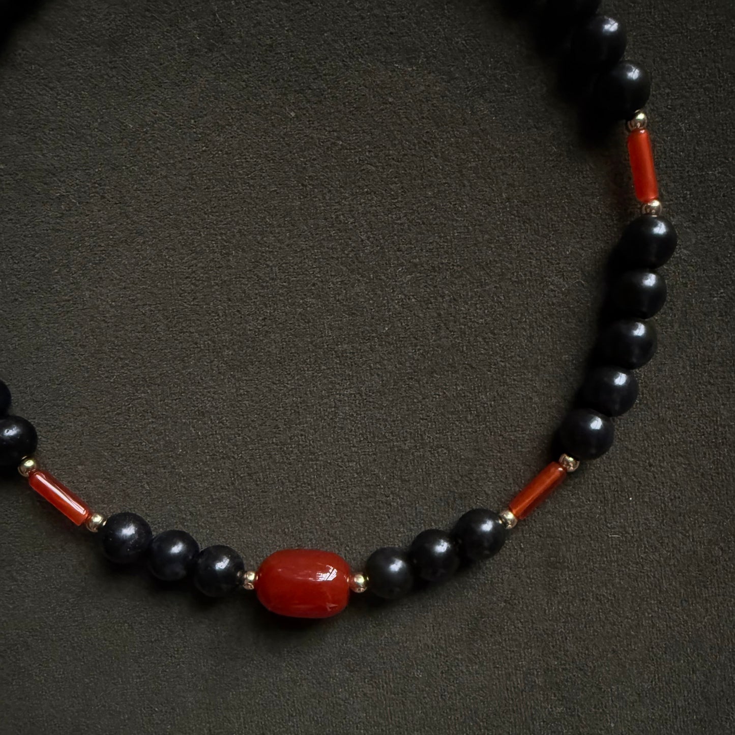 A beaded necklace featuring glossy black round beads, small red cylindrical beads, and a larger polished red oval bead at the center, displayed on a dark textured background.