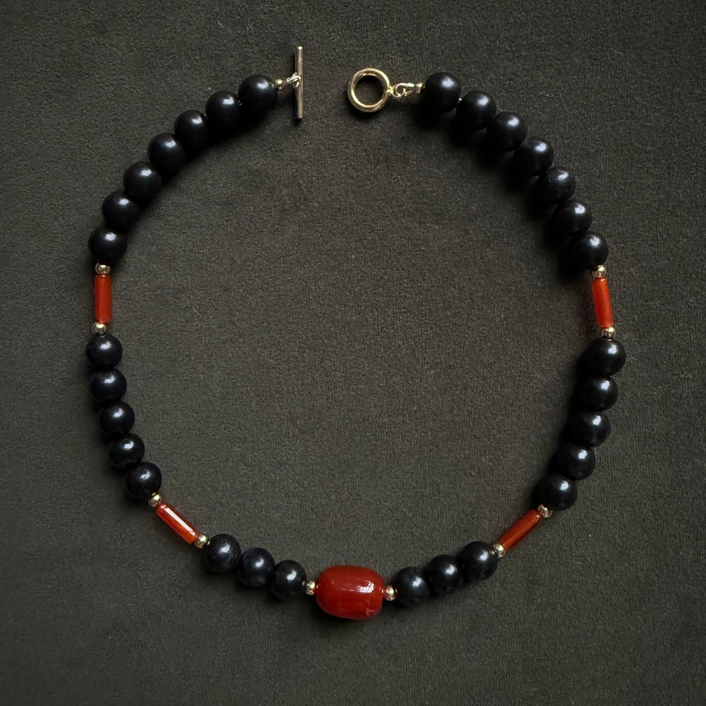 A beaded necklace featuring glossy black round beads, small red cylindrical accents, and a larger polished red oval bead at the center, with a gold-tone toggle clasp, displayed on a dark textured background.