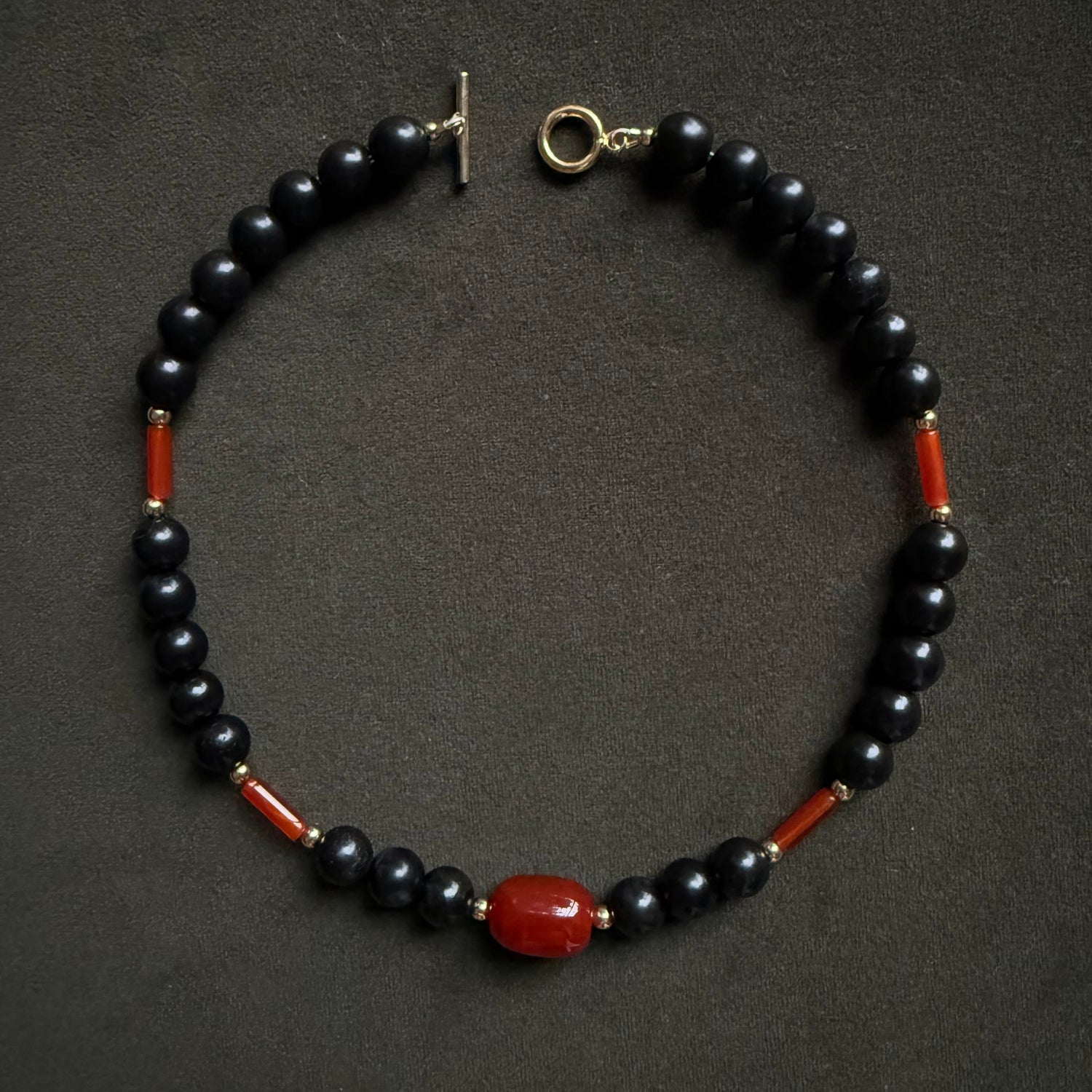 A beaded necklace featuring glossy black round beads, small red cylindrical accents, and a larger polished red oval bead at the center, with a gold-tone toggle clasp, displayed on a dark textured background.