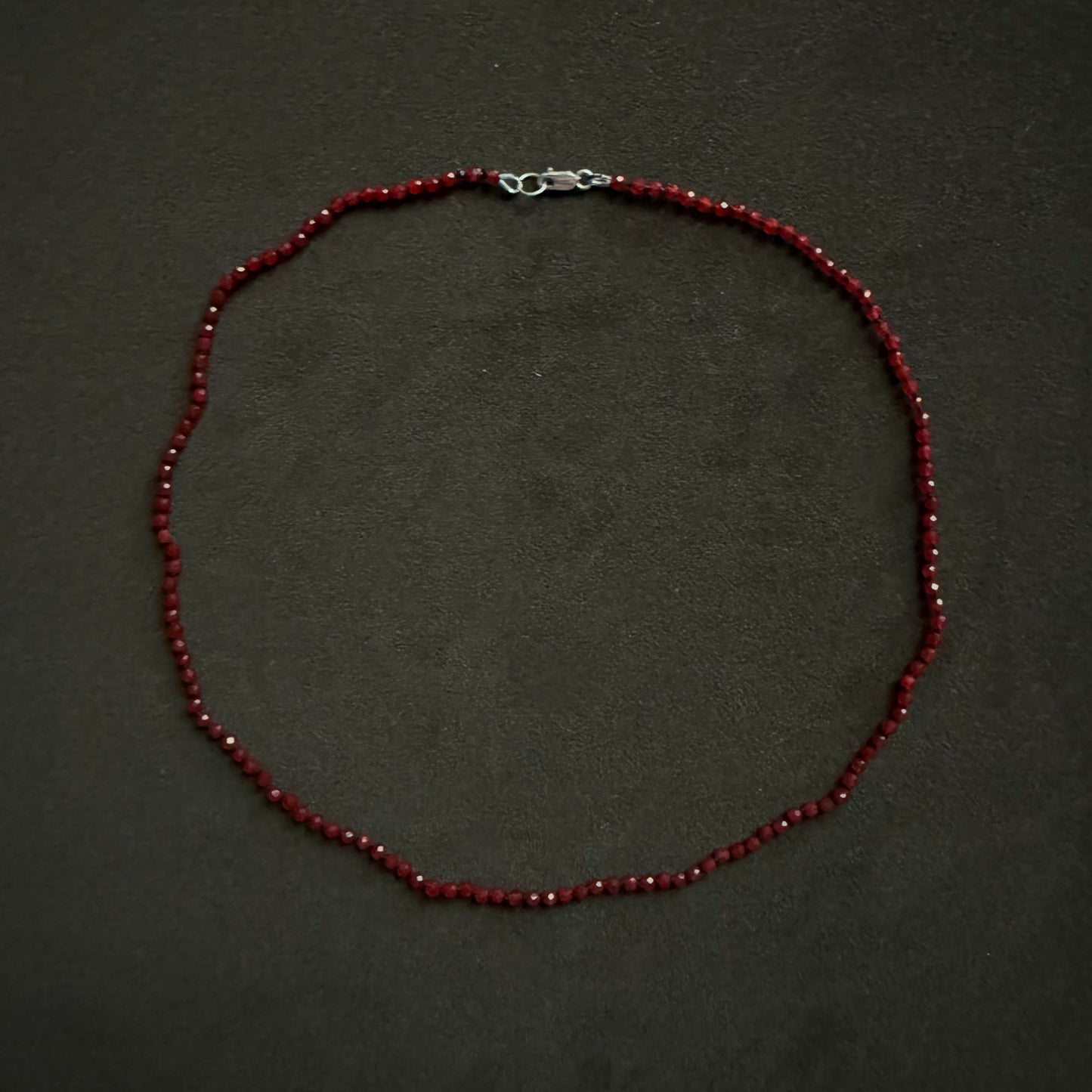 A beaded necklace made of small dark red beads with a silver lobster clasp, displayed on a dark textured background.