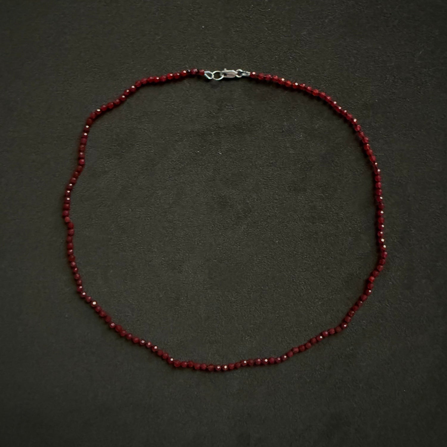 A beaded necklace made of small dark red beads with a silver lobster clasp, displayed on a dark textured background.