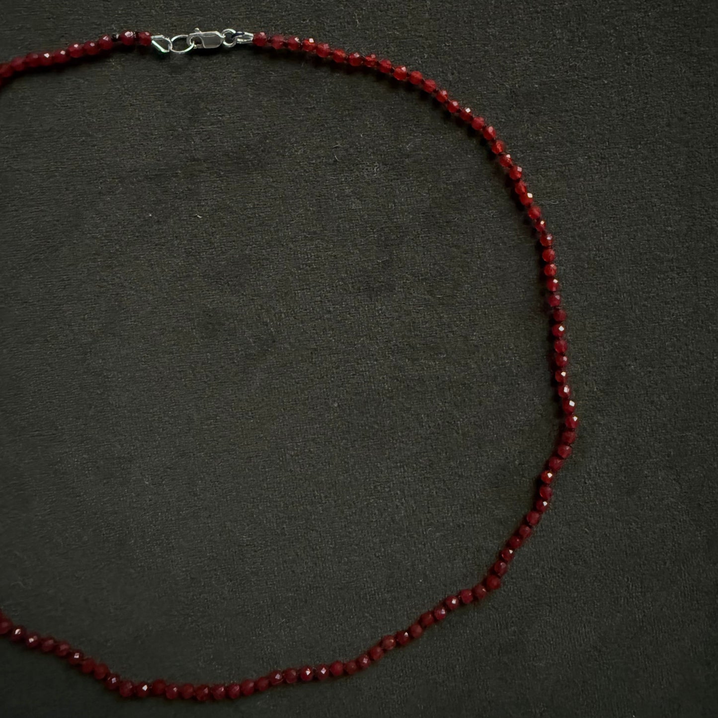 A beaded necklace made of small dark red faceted beads with a silver lobster clasp, displayed on a dark textured background.